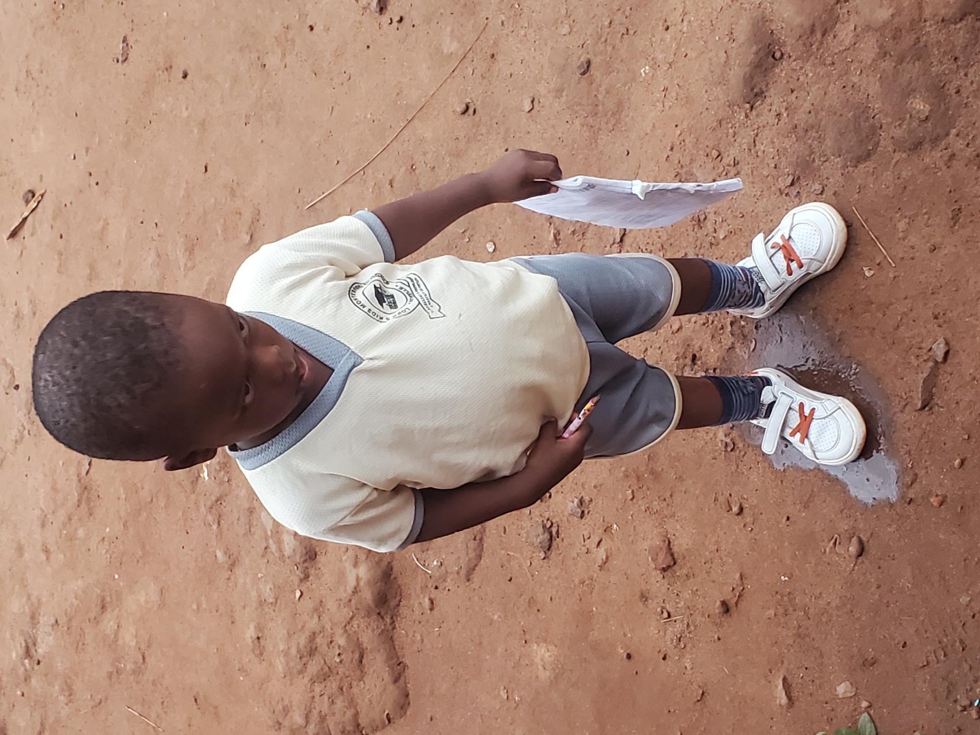 A boy is standing on brown, bare ground in what appears to be Ukwu-Abwa, Nigeria. He is wearing a beige shirt with gray trim and a logo, gray shorts, white sneakers with orange laces, and striped socks. He is holding a piece of paper in one hand and a pencil in the other. One of his sneakers is standing in a puddle.