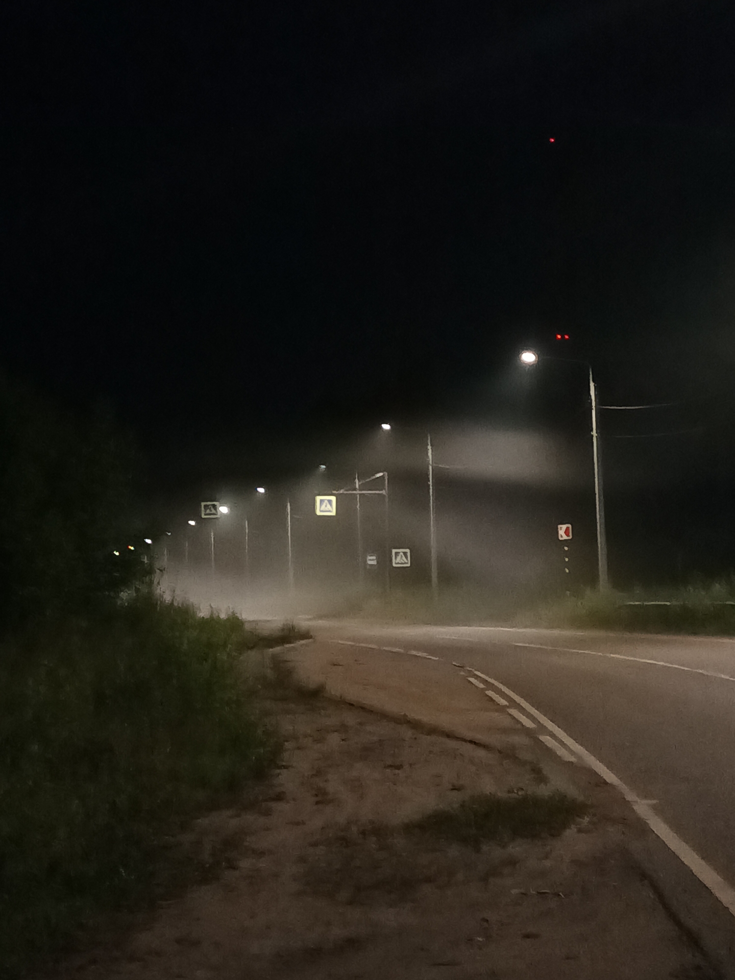 The image depicts a nighttime scene on a rural road in Voskresenskoye, Russia, characterized by heavy fog. A paved road curves to the right, with a double white line marking the center. To the left of the road is a dirt shoulder with sparse grass and vegetation. Further left, a dense line of dark green foliage lines the edge of the road.
Several streetlights line the road, their illumination diffused by the fog, creating hazy glows. Two of these streetlights are equipped with flashing red lights at their tops, and another has a square yellow sign with a pedestrian crossing symbol illuminated. A traffic sign with a red "K" symbol on a white background is also visible on a pole to the right of the road. The sky is dark, with a faint red light visible in the upper portion of the frame, possibly an aircraft warning light. The overall atmosphere is somber and slightly eerie due to the fog and darkness. There are no people or vehicles visible in the image.