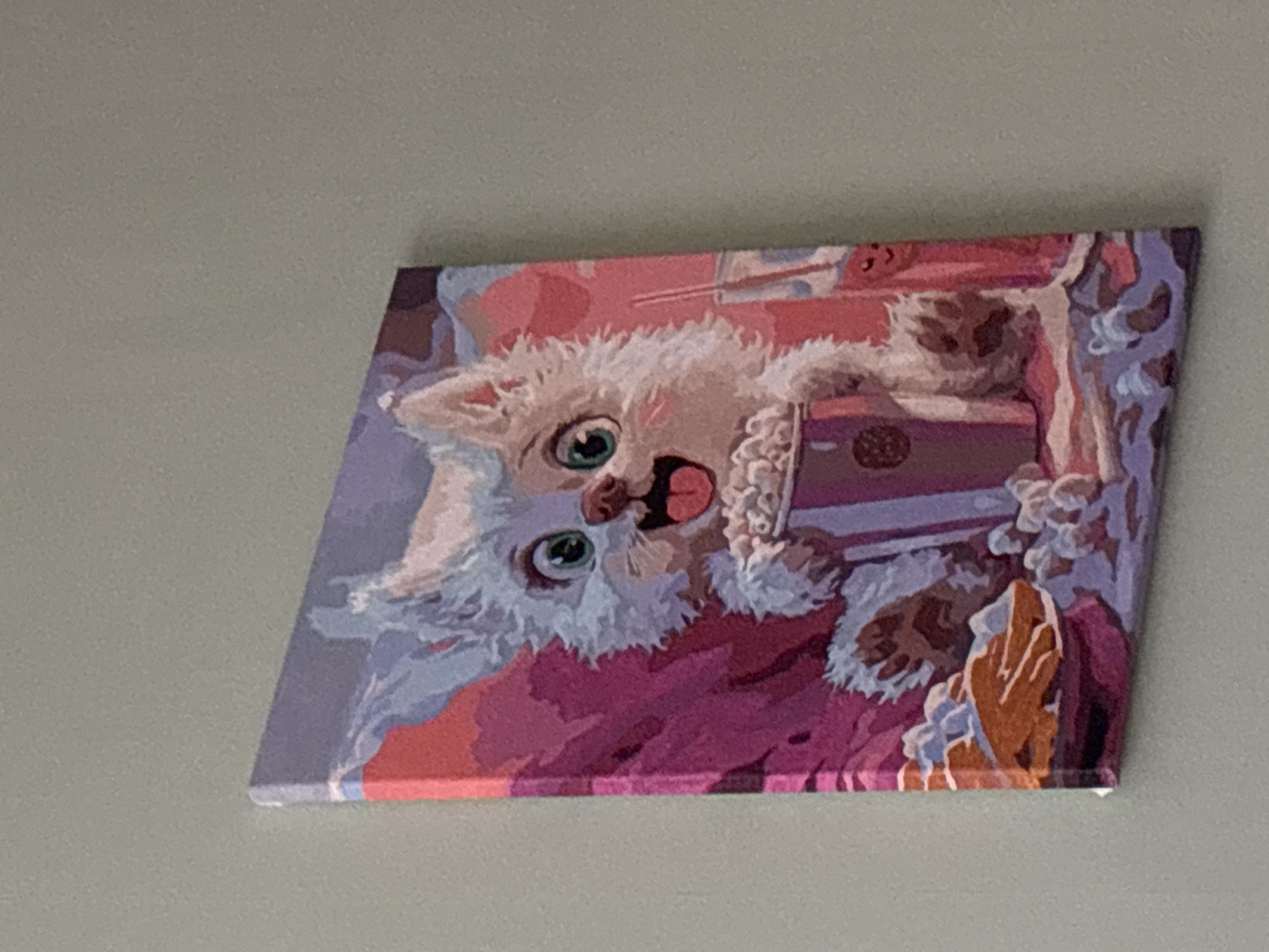 A painting of a white cat with blue eyes, open mouth, pink tongue, and fur around its neck is hanging on a light gray wall. The cat is holding a purple and brown box with a round design on it. The painting has various shades of purple, pink, and orange in the background. The painting is hanging crookedly. The location is Unknown, Russia.