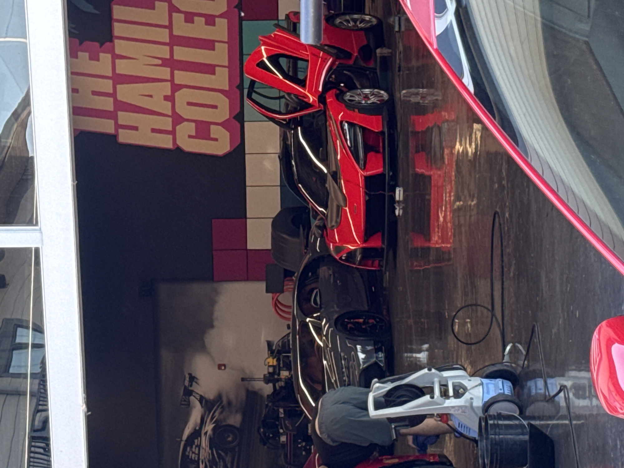 The image captures an indoor scene at "THE HAGMIL COLLECTIO," presumably a car-related business. A bright red sports car stands out, along with other vehicles. A person is present, potentially interacting with a white and blue motorized object. The scene is located indoors in Naperville, United States of America.