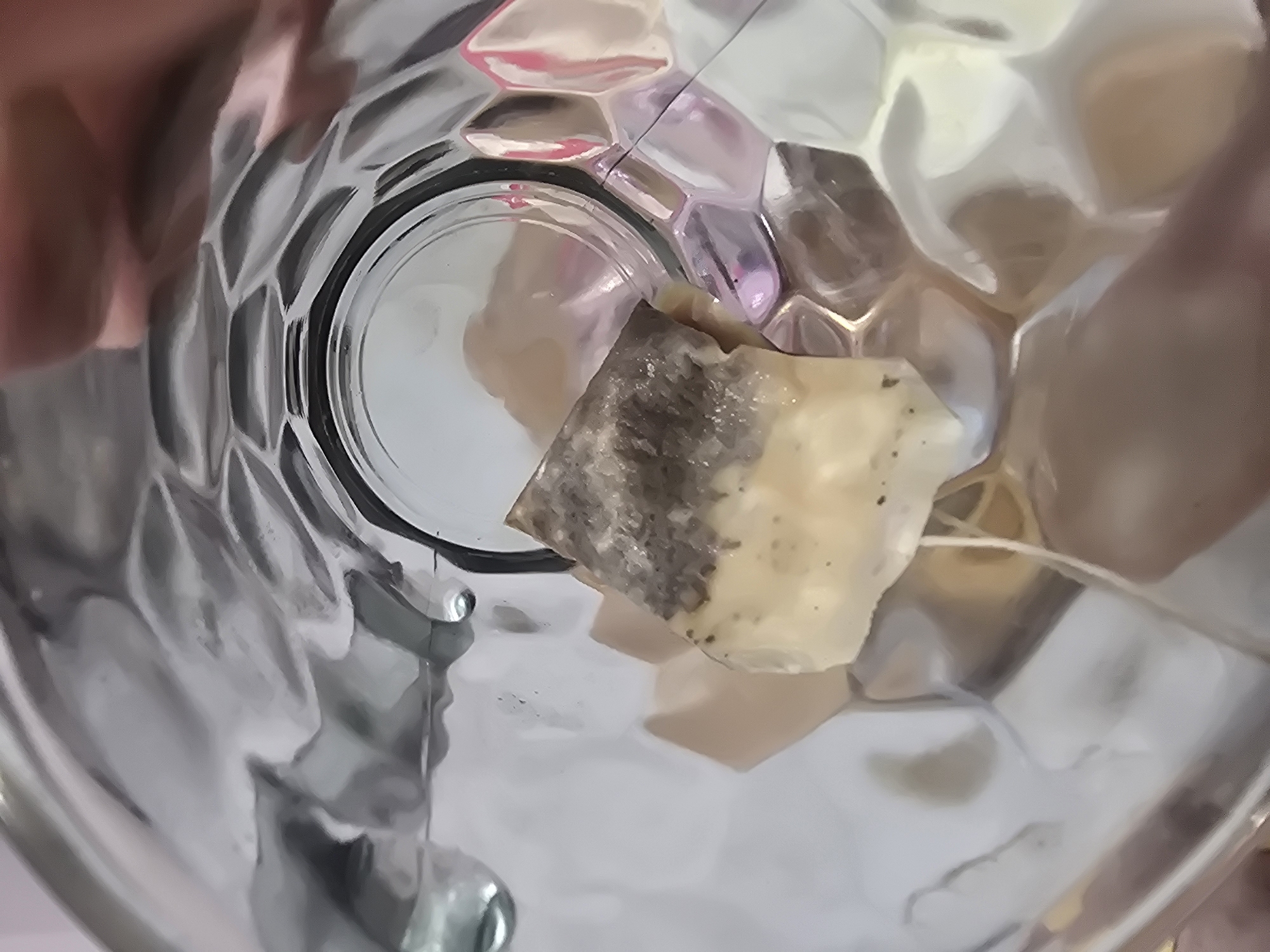 The image is a close-up, overhead shot of a teabag inside a clear glass tumbler. The tumbler has a textured, hexagonal pattern on its exterior, creating reflections and distortions of the light and the teabag. The teabag itself appears to be made of porous paper, filled with tea leaves, and has a white string attached. The background is blurred and consists of abstract shapes and colors, likely reflections within the glass. There is no identifiable information about the location in Ulan-Ude, Russia, or any specific context beyond what is visually present.