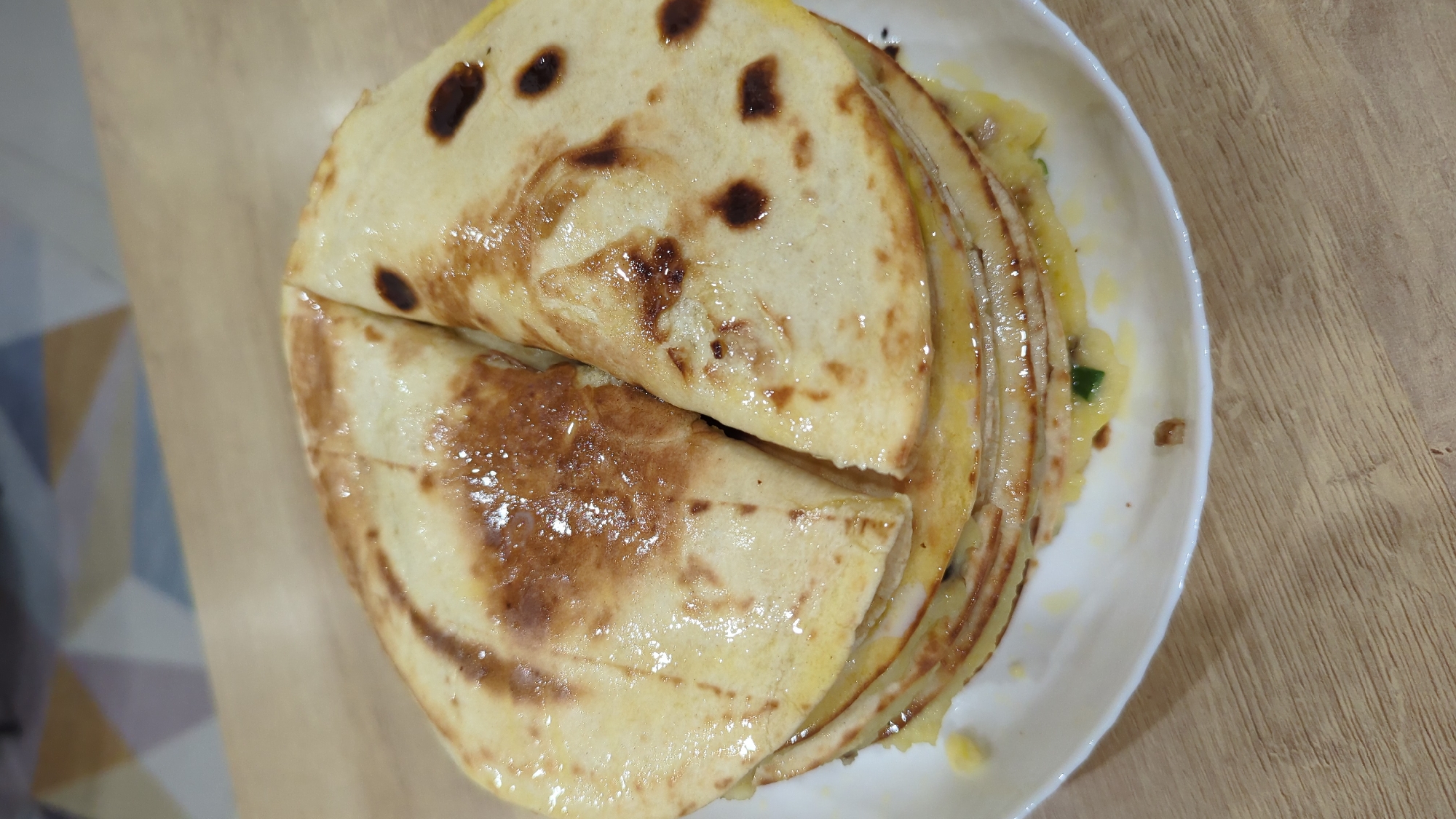 A stack of quesadillas, cut into wedges, is presented on a white plate, which sits on a wooden table. The quesadillas appear to be filled with melted cheese and possibly some green vegetables. The outer tortillas are lightly browned with some darker, seared spots. The lighting suggests an indoor setting, possibly a kitchen or dining area. There is no indication of the specific location in Bac Nha Trang Ward, Vietnam. The scene is static, with no people or ongoing actions depicted.
