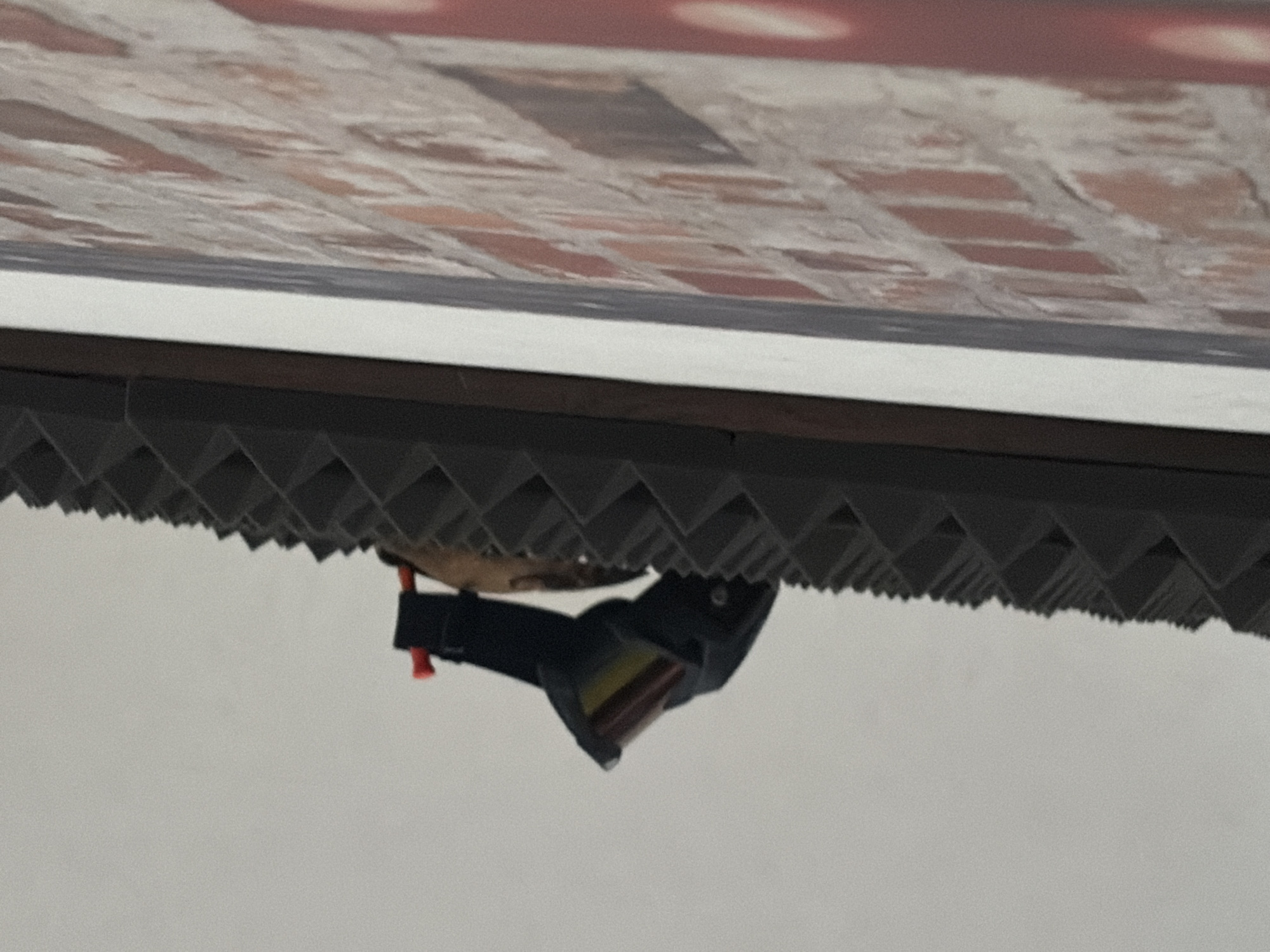 A pair of dark-colored goggles with a red clip is hanging from a textured surface. The textured surface is dark gray and has a repeating pyramidal pattern. To the right of the textured surface is a white border, and to the right of that is a brick wall. The brick wall is composed of orange and red bricks with gray mortar. The image is taken from a slightly upward angle. The background is a plain, light gray. The scene is indoors, likely a studio or a room with acoustic paneling.