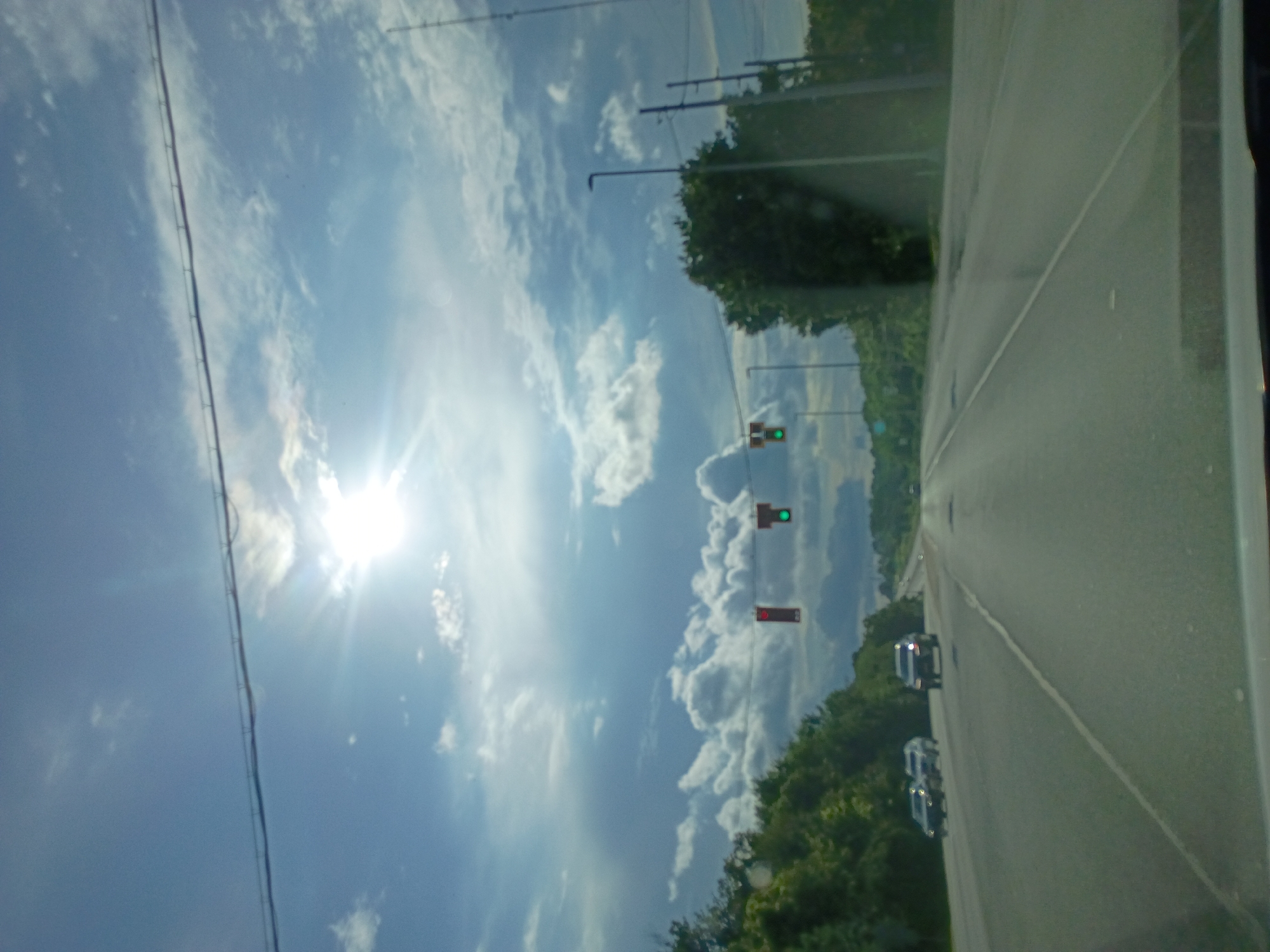 The image shows a view from inside a vehicle traveling on a multi-lane highway, likely in Versailles, United States. The sun is bright and high in the sky, with scattered clouds. Traffic lights are visible suspended over the highway, with one showing a red light and another showing a green light. Cars are traveling in the same direction as the viewer. The sides of the highway are lined with trees and vegetation. The road surface appears to be asphalt, with clear lane markings. Utility poles and streetlights are also present along the roadside. The overall scene depicts a typical highway environment on a sunny day.