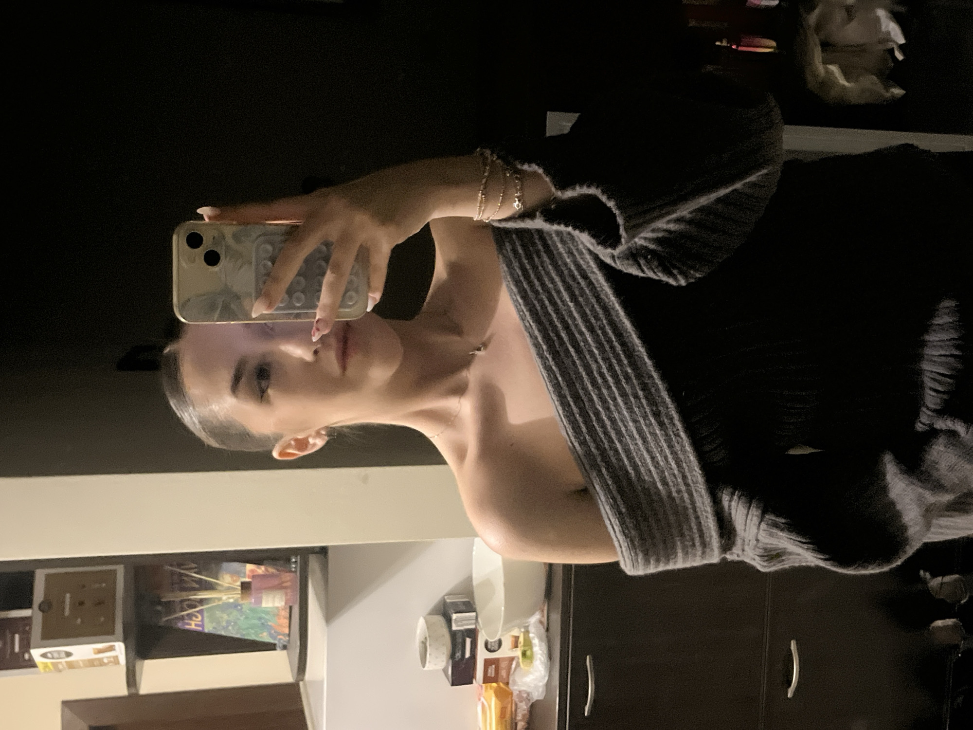A person is taking a mirror selfie with a smartphone, the frame of the phone is gold. The person has on a grey, oversized cardigan, with layered bracelets on their wrist. They are wearing a necklace. The background appears to be in a house, near a dresser in the kitchen or bathroom. The location cannot be determined to be Sofia, Bulgaria from the image provided.