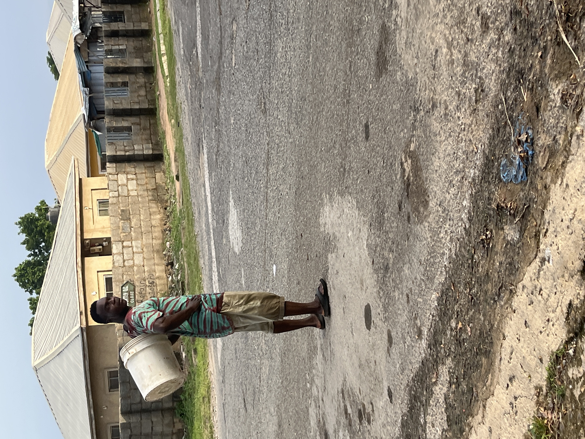 In Abuja, Nigeria, a man stands on a paved road carrying a large white bucket on his right shoulder. He is wearing a short-sleeved shirt with horizontal green and maroon stripes, and khaki shorts that end just below his knees. His feet are bare except for black flip-flops. Behind him, a low stone wall separates the road from a residential area with houses featuring corrugated metal roofs and block construction. The environment appears to be an urban or suburban street, with some greenery visible along the roadside and scattered debris on the pavement. The sky is overcast.
