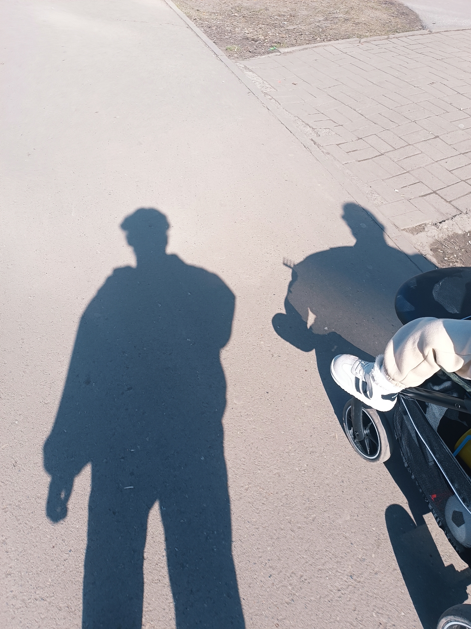 The image shows an aerial view of a person pushing a stroller on a paved surface, likely a sidewalk or path. The person's shadow is visible, cast to the left of the frame. The stroller contains a child whose leg and foot, wearing a white Adidas sneaker, are visible. The stroller has a black frame and black mesh storage underneath. The surface is partly concrete, with a section of paved blocks to the upper right. The location is Izhevsk, Russia.
