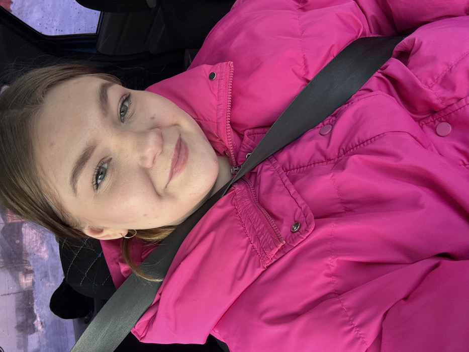 The image depicts a young female inside a vehicle, located in the city of Фоки, Russia. The subject, a Caucasian female with light brown hair and light-colored eyes, is visible from the chest up, occupying either the front passenger or driver's seat. She is wearing a bright pink padded winter jacket, which features a visible zipper closure and multiple snap buttons, some pink and some dark-colored. A dark gray seatbelt is fastened across her torso, extending diagonally from her left shoulder to her right hip. Her hair is partially pulled back, and she wears small, light-colored hoop earrings. She exhibits a subtle smile, looking directly into the camera. The background, visible through a vehicle window on the right side, shows a blurred outdoor scene with pale, possibly snowy conditions and indistinct distant elements. The interior of the vehicle appears dark-toned, with a dark headrest visible behind her left shoulder. The lighting is diffuse, suggesting an overcast daytime environment.