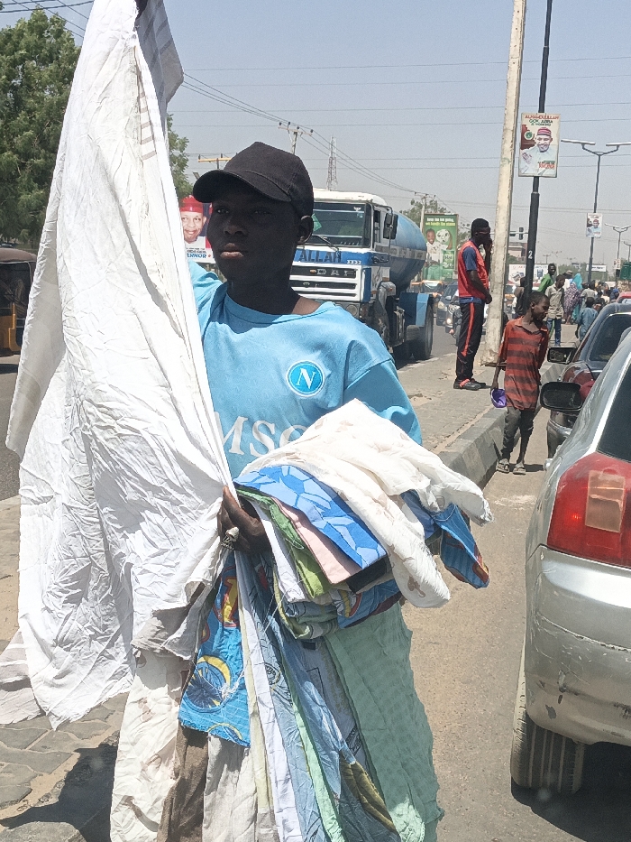 This image captures a street scene in Hotoro South, Nigeria, under bright daylight. In the immediate foreground, a young male, appearing to be a vendor, with dark skin and a neutral expression, stands facing the camera. He wears a black baseball cap and a light blue short-sleeved t-shirt with a white "N" logo on the left chest and "MSC" text below it. He holds a large, crumpled bundle of white fabric in his left arm, extending upwards. In his right hand, he grasps a stack of folded, multicolored textiles, likely sheets or fabrics, which extend downwards to his side. A ring is visible on his right ring finger.
To his immediate right, the rear portion of a silver sedan is visible, parked or moving slowly. Behind the primary subject, a white DAF truck, possibly a cement mixer with a blue drum and "ALLAH" written on its front, is present in the midground. Further back and to the left, part of a yellow auto-rickshaw (keke Napep) is discernible. A political poster, partially obscured, depicting a man in a red cap with "GOVERNOR" text, is visible behind the vendor's left shoulder.
In the midground to the right, a male in a red and black tracksuit stands by a utility pole, appearing to be on a mobile phone. A young child in a red and black striped t-shirt and dark pants stands nearby, holding a purple object. The street beyond is populated with other pedestrians and vehicles, including other cars and motorcycles. A second political poster featuring a man in a white cap is affixed to the utility pole. The sky is clear and bright, indicating a sunny day. The scene depicts a bustling roadside environment.