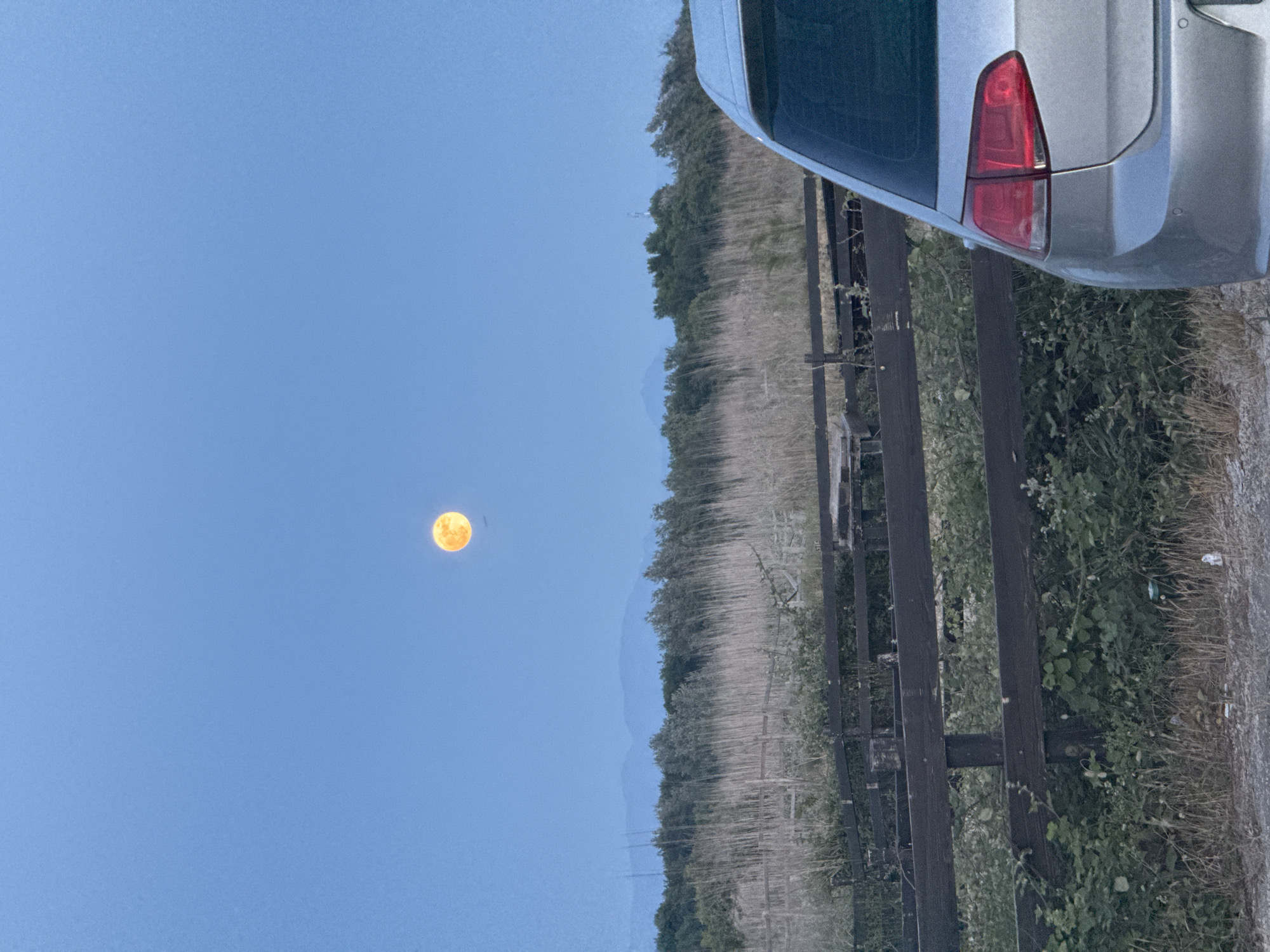 The image shows a landscape in Gornji Štoj - Shtoji i Epërm, Montenegro at twilight. The sky is a pale blue, and a full, orange moon is visible. A silver car is parked on the right, partially obscuring a field of tall, dry grass and green vegetation. A dark wooden fence runs along the edge of the field. In the distance, a line of trees and low mountains can be seen. The image is rotated counterclockwise.