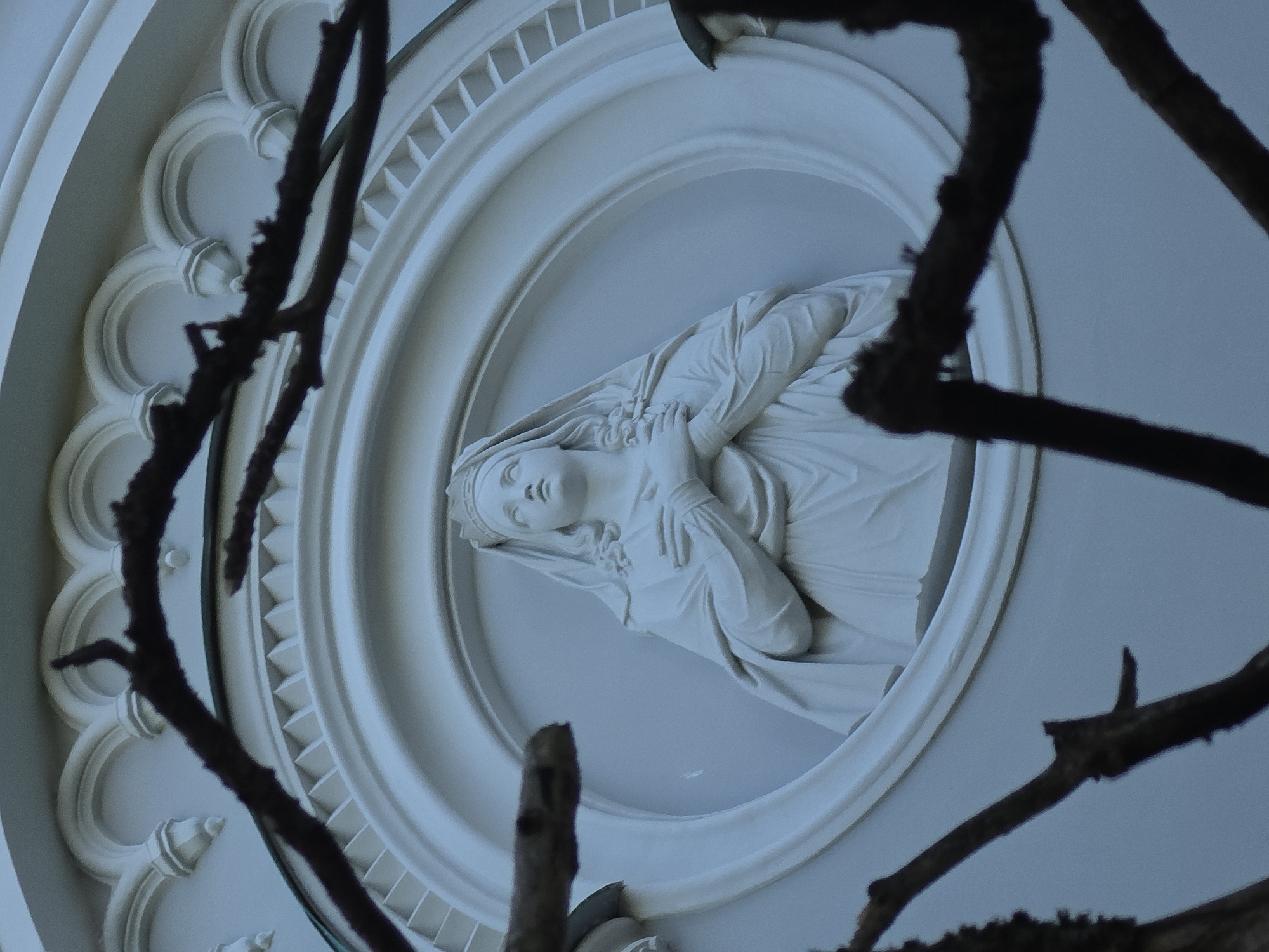 The image shows a relief sculpture of a woman with a crown and veil, possibly the Virgin Mary, set within an oval frame on the exterior of a white building. The sculpture is detailed, with the woman's hands crossed over her chest and her head tilted slightly upwards. The building's facade features architectural details such as arched decorative elements above the oval frame and a patterned band below it. Dark, bare tree branches obscure parts of the sculpture and the building, creating a somber or perhaps seasonal atmosphere. This sculpture is located on the north facade of the Pavlovsk Palace in Gatchina, Russia.