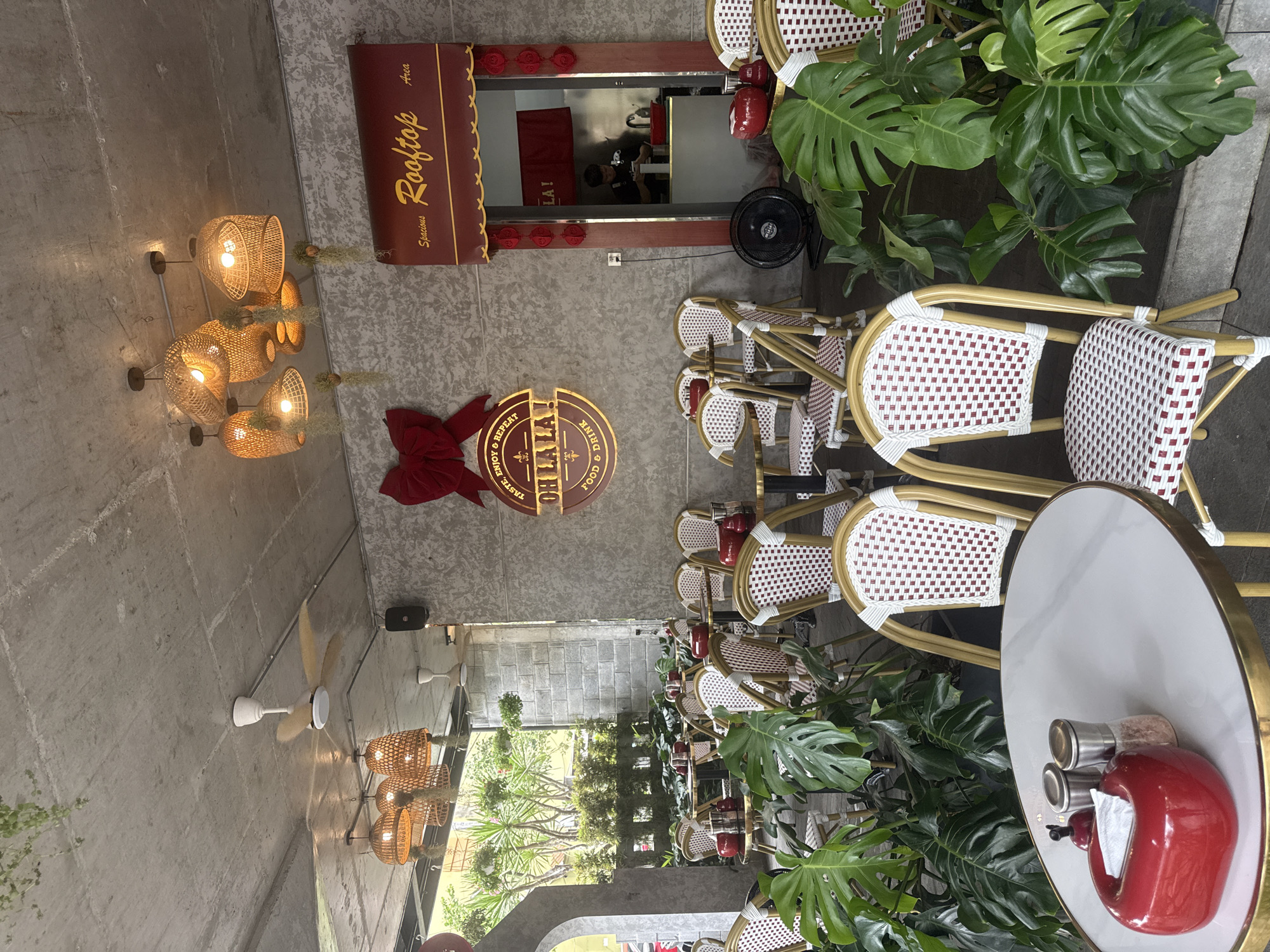 The scene depicts an outdoor dining area with a modern, plant-filled aesthetic. Numerous wicker chairs with white and red patterned seats are neatly arranged around small, round tables. Some tables are set with red teapots, salt and pepper shakers, and napkins.
The environment is accented with lush green plants, including large Monstera leaves, which add a touch of nature to the space. A decorative sign reading "OH LA LA FOOD & DRINK" is visible on the wall, along with a "Rooftop Area" sign above a service window.
Lighting fixtures with a woven design hang from the ceiling. The setting suggests a stylish, urban café or restaurant.