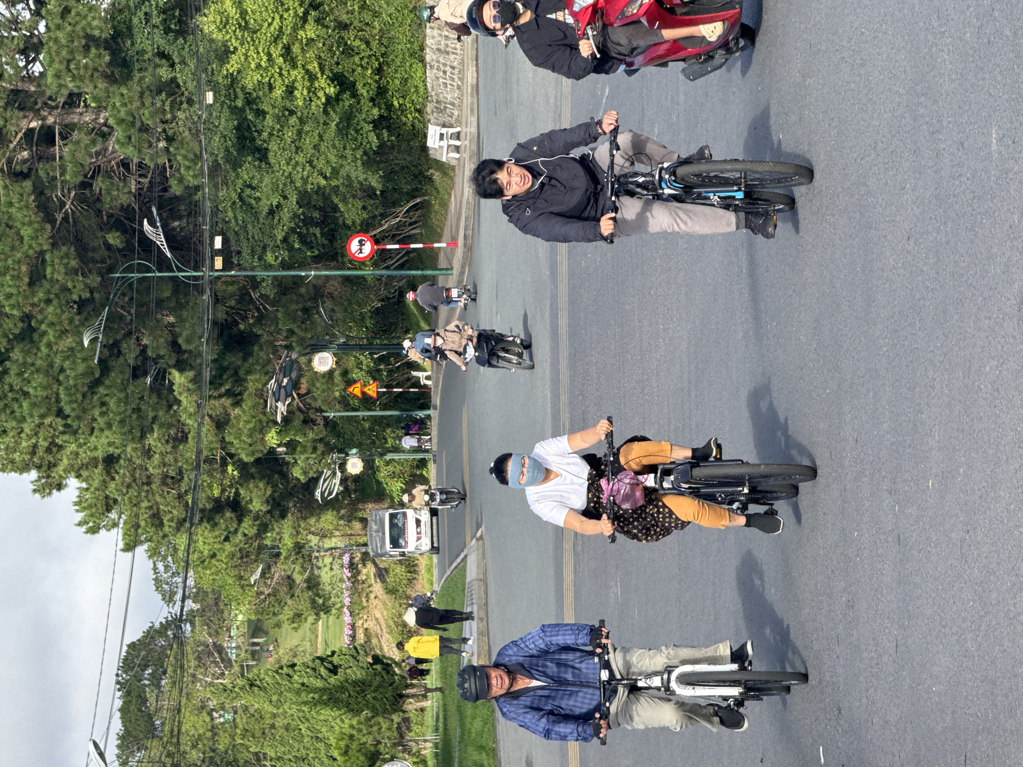 The image captures a street scene in Phường Xuân Hương - Đà Lạt, Vietnam, on a bright, overcast day. The road is paved with asphalt and marked with yellow lines. In the foreground, four individuals are riding bicycles and motorbikes.
On the left, an elderly man with a blue plaid jacket, light-colored pants, and a black helmet is riding a white bicycle. To his right, a woman wearing a white t-shirt, patterned skirt, and orange pants, with a light blue mask covering her lower face, is also riding a bicycle. Further to the right, a man in a black jacket and grey pants is on a black bicycle. To the far right, partially visible, is a person on a red motorbike wearing a black jacket and a face mask.
In the background, several other individuals are seen on motorbikes and on foot. A white delivery truck is parked on the right side of the road. The street is lined with lush green trees and some flowering bushes. Streetlights and electrical wires are visible overhead. In the distance, a gentle slope with more trees and some buildings suggests the hilly terrain of Đà Lạt. Several traffic signs are posted along the roadside, including a circular red and white sign with a horn symbol crossed out, indicating no honking. The overall atmosphere appears to be one of daily activity and commuting within a scenic urban environment.