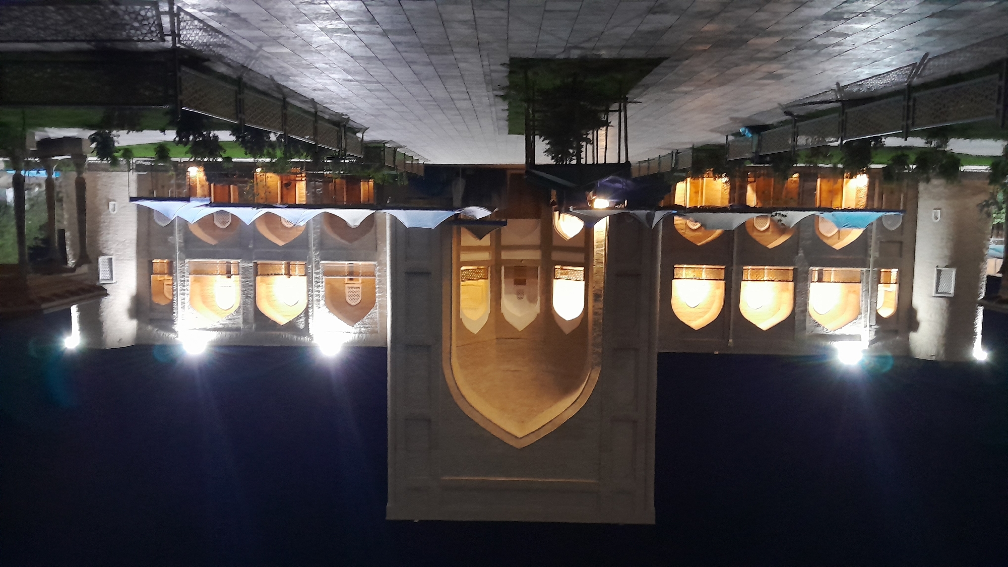 The image depicts an inverted view of a building at night. The building features ornate architectural details, including arched windows with intricate lattice work and a large, shield-shaped central facade. The structure is illuminated by warm, yellow lights emanating from within the windows, creating a welcoming glow. The foreground is dominated by a dark, reflective surface, possibly water or a polished floor, mirroring the lights and architectural elements. Beyond the building, a tiled walkway is visible, with trees and a decorative fence lining the perimeter. The overall scene suggests a public space or a cultural landmark, possibly a mosque or a historic building, in Denov, Uzbekistan. The time of day is clearly night due to the darkness and artificial lighting. The weather appears calm, with no indications of rain or wind. The architecture and lighting convey a sense of grandeur and cultural heritage. No people are visible in the image.