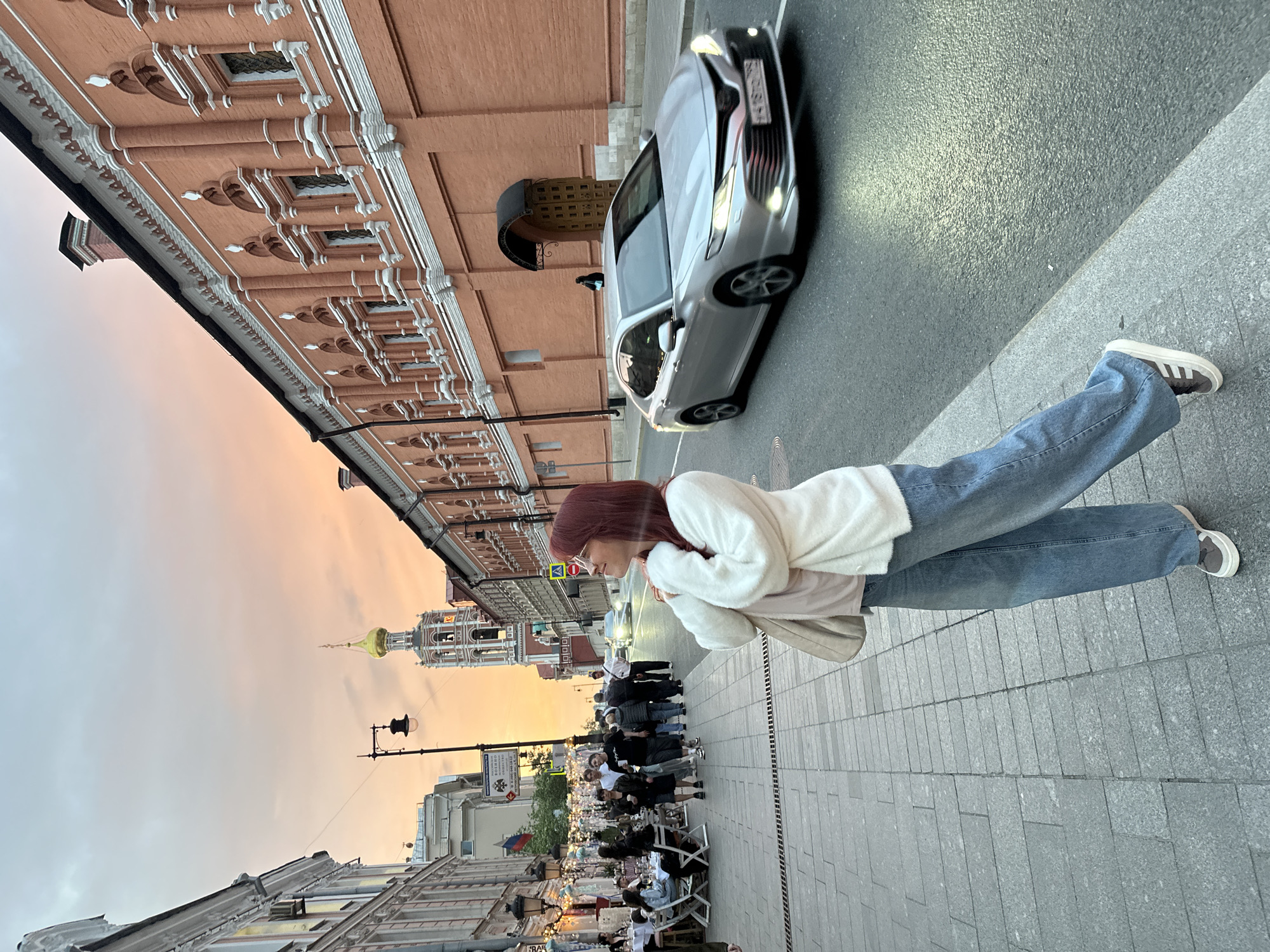 The image captures a scene in an urban environment. A woman stands on a paved sidewalk, wearing jeans, a white jacket, and sneakers. A silver car is parked on the adjacent street. In the background, a building with ornate architectural details stands out, along with a tower crowned with a golden dome. A group of people is lined up on the sidewalk, and beyond them, tables and chairs suggest an outdoor dining area. The sky displays colors of sunset or dusk.
