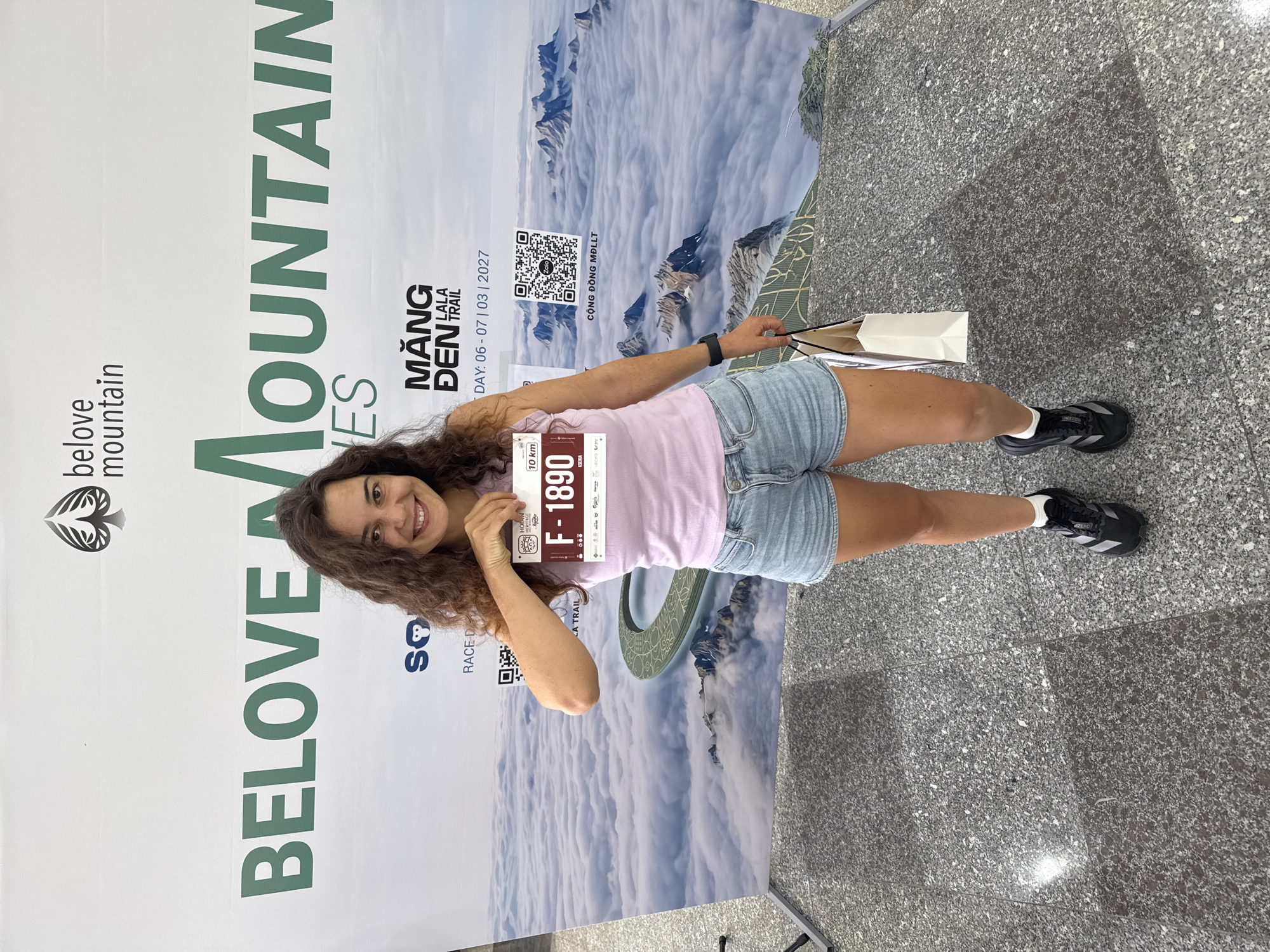 A smiling woman stands in front of a large banner for "BELOVE MOUNTAIN" and "MANG DEN LALA TRAIL" in Hoi An, Vietnam. The banner features a scenic background of mountains shrouded in clouds and a circular path. The woman, with curly brown hair, is wearing a light pink tank top, denim shorts, and black athletic shoes. She is holding up a race bib with the number "F-1890" and "10 km" visible. She is also carrying a white shopping bag in her right hand. The floor is made of speckled granite tiles.