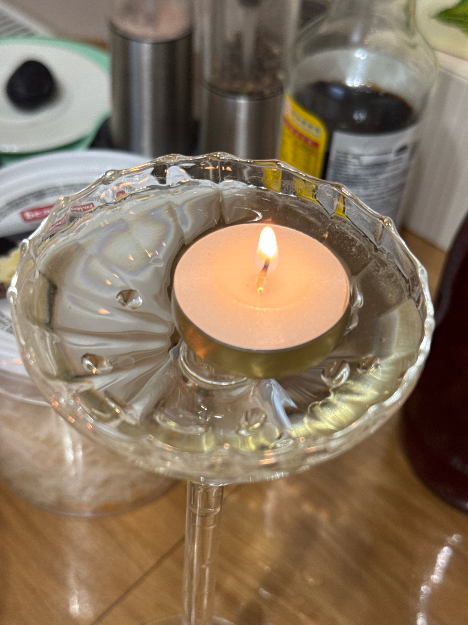 The image shows a lit tea light candle in a decorative glass holder. The candle flame is bright orange. The glass holder is clear with intricate patterns and appears to be filled with water. In the background, there are various objects, including what looks like a spice grinder, a bottle of dark liquid (possibly soy sauce or balsamic vinegar), and a white plate with a dark spherical object on it. The scene is set on a wooden surface. There is no indication of the location being in Захарьино, Russia, or any other specific city.