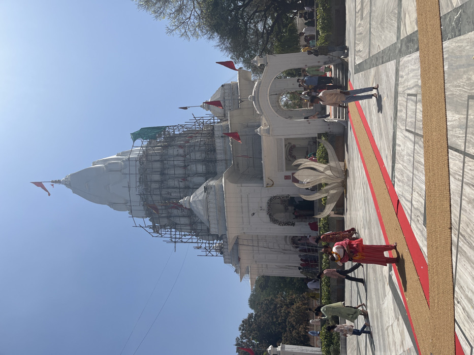 The image shows a white temple under construction, located in Panchkula, India (latitude: 30.725, longitude: 76.856). Scaffolding is erected around the temple, and a red flag flies from the top. A red carpet leads to the temple entrance, and several people are standing around the courtyard. A woman carrying a child walks toward the temple along the carpet. There are trees and green grass in the background. The sky is clear and blue. The temple is surrounded by a marble-like pavement. The courtyard is clean and well-maintained.