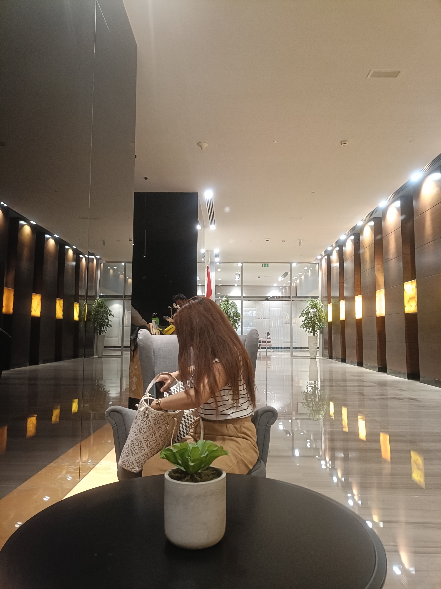 The image depicts a modern indoor space, likely a hotel lobby or a high-end commercial area in Dubai, United Arab Emirates, characterized by sleek design elements and ambient lighting. A woman with long, dark brown hair, wearing a striped top and a tan skirt, is seated in a plush grey armchair, facing away from the camera. She is holding a smartphone in her right hand and has a patterned tote bag resting on her lap. To her right, on a black circular table, sits a small potted plant with green leaves in a grey concrete pot.
The background reveals a long, polished corridor with a highly reflective marble floor. The walls are adorned with vertical wooden panels interspersed with illuminated rectangular sections in warm yellow hues, creating a sophisticated and inviting atmosphere. The corridor stretches into the distance, leading to glass-enclosed offices or meeting rooms with visible interior lights. Two large potted plants with lush green foliage are strategically placed along the corridor, adding a touch of nature to the urban setting. The ceiling is a neutral color with recessed lighting and ventilation fixtures. The overall impression is one of contemporary luxury and spaciousness, typical of establishments in Dubai.