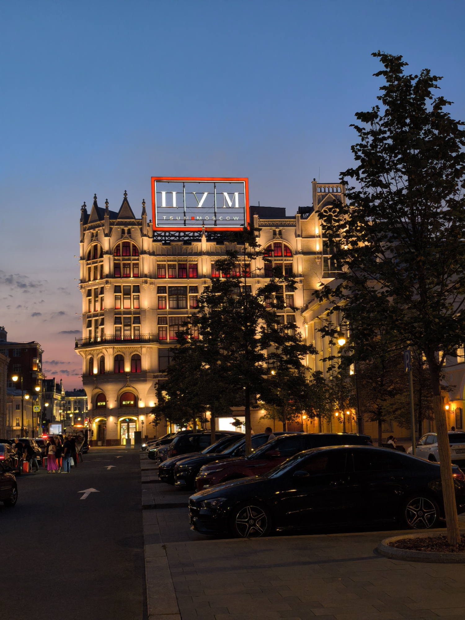 The image shows a building in Moscow, Russia, at dusk. The building is lit up and has a sign that says "TSUM MOSCOW". There are cars parked on the street in front of the building. People are walking on the sidewalk. The sky is a deep blue color. The scene is illuminated by streetlights and the lights from the building.