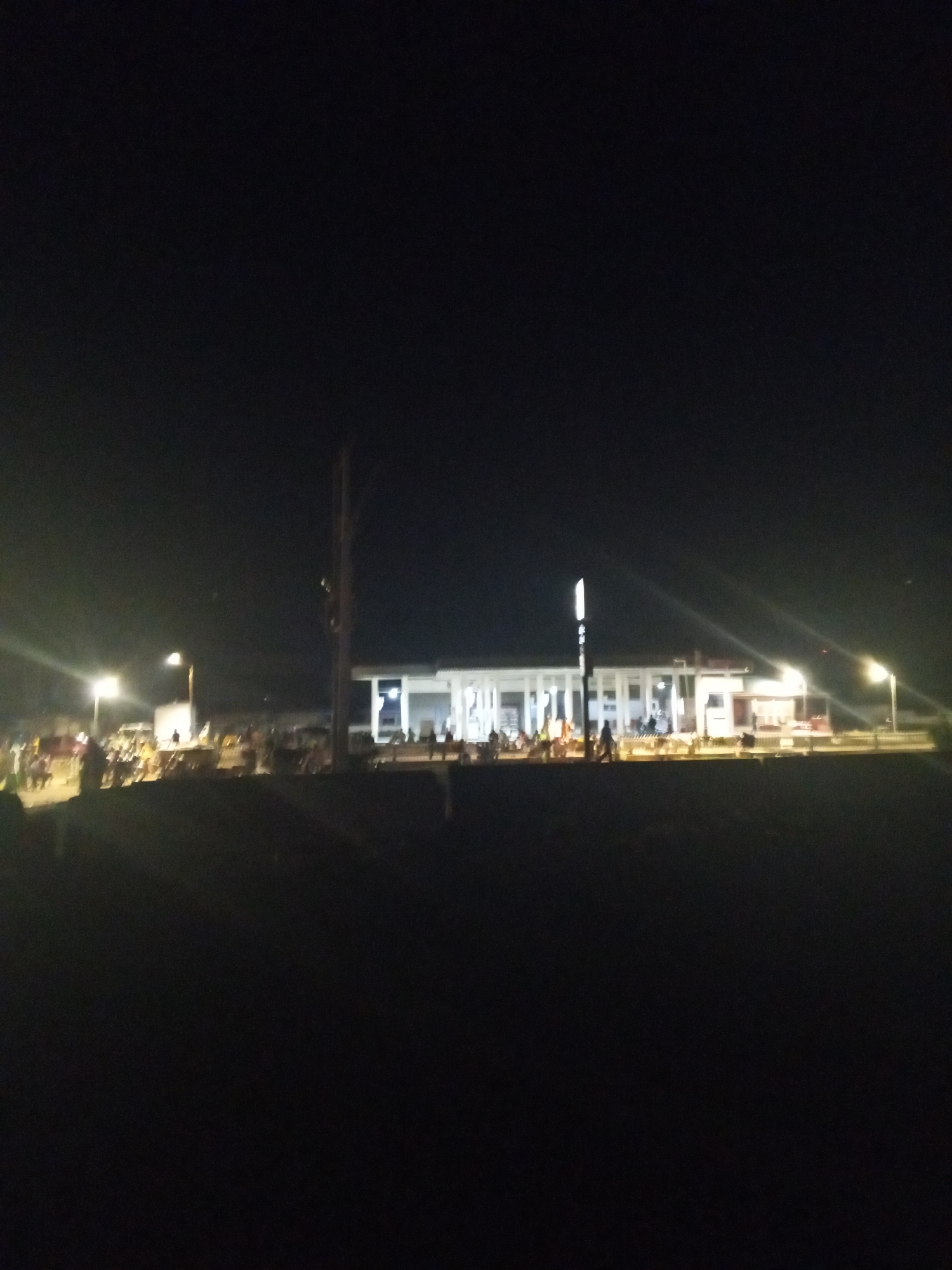At night in Lokoja, Nigeria, a well-lit, white building stands in the background. Groups of people are gathered in front of the building, illuminated by streetlights. The foreground is dark, obscuring details of the ground. A tall, thin pole is positioned between the crowd and the building.