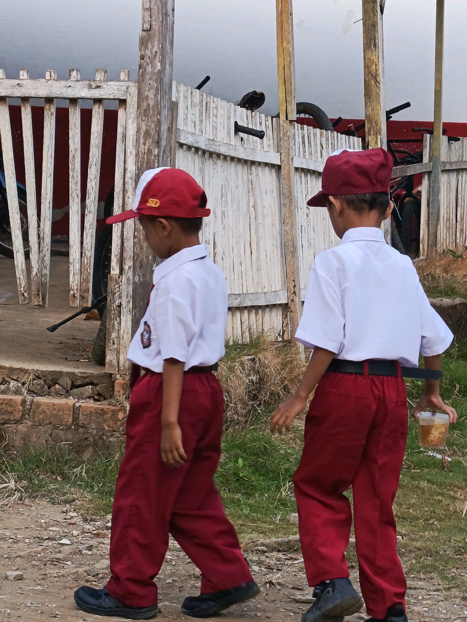 Two young boys are walking on a dirt path in front of a weathered wooden fence. The boy on the left is wearing a white short-sleeved shirt with a red collar and trim, a red baseball cap with a white top, and red pants. He has a patch on his left sleeve. The boy on the right is wearing a similar uniform: a white short-sleeved shirt, a red baseball cap, and red pants. He is holding a clear plastic cup with a light brown beverage in his right hand. Behind the fence, bicycles are leaning against a white wall. The environment appears to be outdoors, with dry grass and some brick debris on the ground. The overall scene suggests a rural or semi-urban setting in Megang Sakti II, Indonesia.