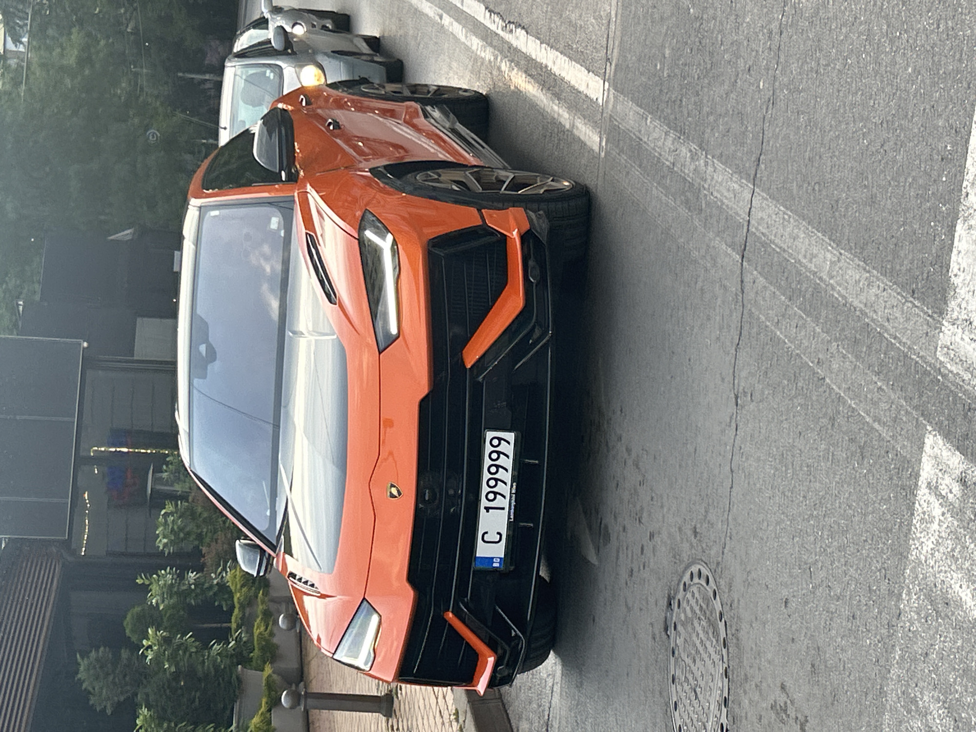An orange Lamborghini Urus SUV is centered in the foreground, facing the viewer and appearing stationary in a line of traffic on an asphalt road. The vehicle features a contrasting black hood insert, black grille, and bronze-colored multi-spoke wheels. Its front license plate displays "BG C 199999" with "Lamborghini Sofia" inscribed below the numbers, indicating its registration in Sofia, Bulgaria. A blurred reflection of a person, likely the driver, is visible through the windshield.
Behind the Lamborghini, a silver hatchback is visible, followed by another light-colored vehicle, also appearing to be in traffic. The road surface includes a circular metal manhole cover in the lower left and faded white painted lines on the right, possibly part of a crosswalk or lane demarcation.
To the left, a dark grey bollard stands on a paved sidewalk. Behind it are large planters containing green bushes and small trees. A dark building with reflective windows and visible interior lighting or reflections is situated behind the foliage. On the right side of the background, lush green trees indicate a vegetated area. The scene is illuminated by soft, diffused light, suggesting late afternoon or early evening.