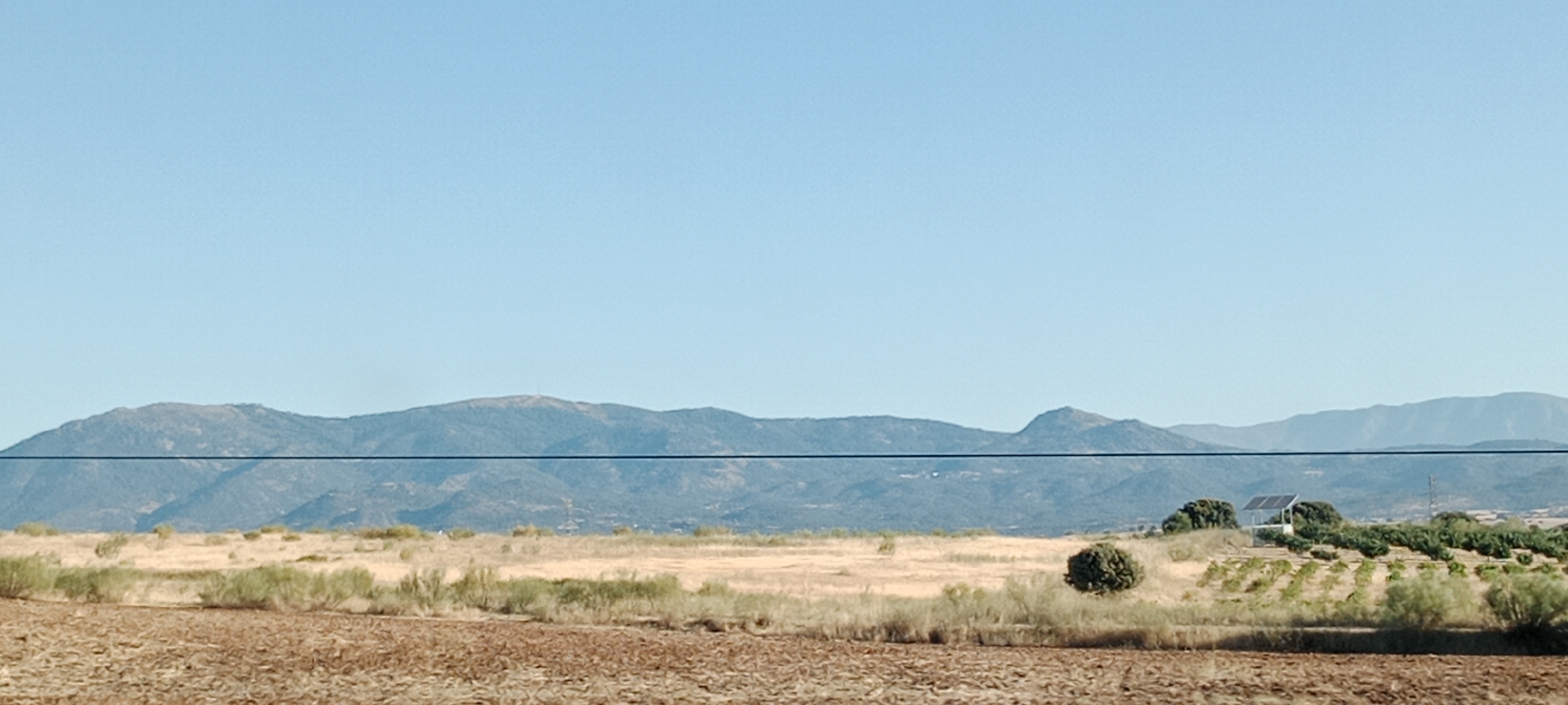 This image depicts a rural landscape with a clear blue sky. In the foreground, a tilled field of reddish-brown soil is visible. Beyond this field, a dry, grassy plain stretches out, dotted with sparse green shrubs and trees. A thin black horizontal line, likely a wire, traverses the middle of the image. In the mid-ground, a cluster of trees and rows of young plants are present, with a small building and solar panels situated on a slight elevation. The background is dominated by a range of low, rolling mountains covered in muted green vegetation under the vast expanse of the sky. The scene is likely located in the municipality of Lucillos, Spain, given its characteristic dry, agricultural landscape and mountainous horizon. No people or animals are visible in the frame.