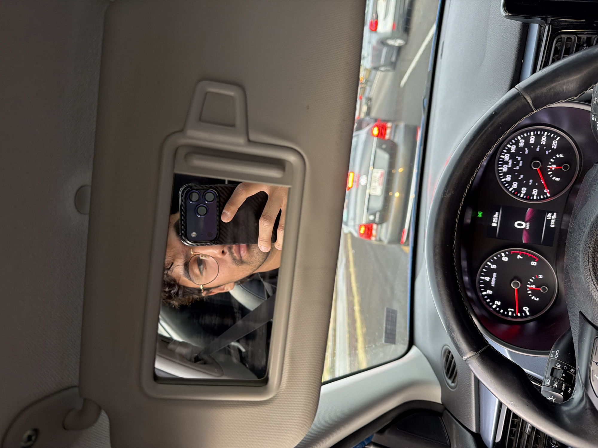 The image shows the interior of a car in heavy traffic, likely in San Bernardino Chalchihuapan, Mexico, as indicated by the OCR text reading "253m", which could represent a local road or landmark. A man's reflection is visible in the sun visor mirror, indicating he is taking a selfie with his phone. The dashboard features a speedometer reading 0 km/h and a temperature of 18°C. The mileage is 67415 km. The steering wheel is black. Outside the car, traffic is congested with several vehicles visible.