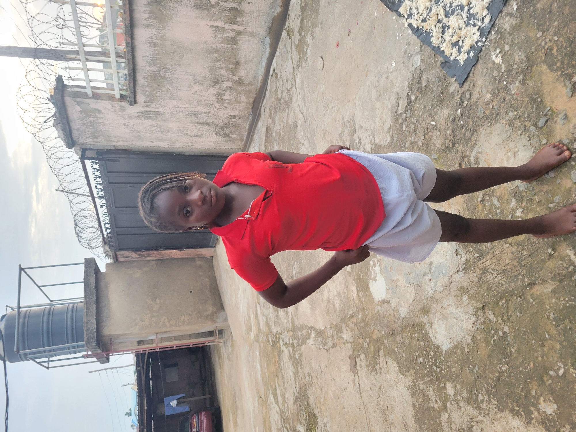 A young girl stands facing forward in an outdoor courtyard, with her hands on her hips. She is wearing a red polo shirt and white shorts. Her hair is styled in braids. Behind her is a large black water tank on a concrete pedestal, a black metal gate topped with barbed wire, and a light-colored wall. To the left, a red car is partially visible, and laundry is hanging on a line. The ground is paved with concrete and shows signs of wear and some dampness. In the lower right corner, a black tarp is laid out with small, white items scattered on it. The sky is overcast. The scene appears to be in Madalla, Nigeria.