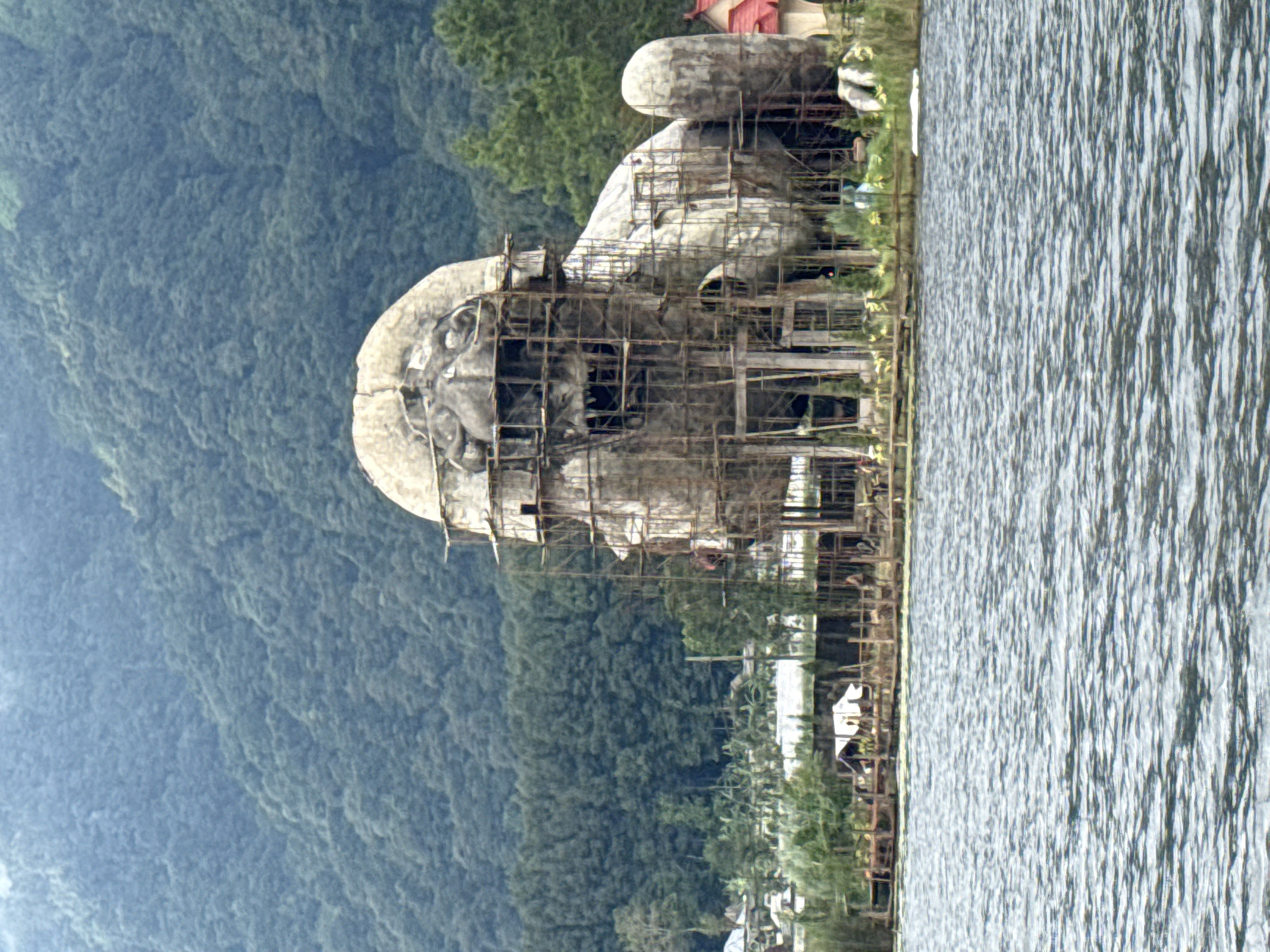 The image depicts a large statue of Buddha, partially constructed with scaffolding, located near Lake Beratan in Baturiti, Indonesia. The statue is gray and appears to be made of stone or concrete. It features a visible facial structure. A network of scaffolding surrounds the statue. Lush, green vegetation covers the hillside in the background. The foreground shows the waters of Lake Beratan.