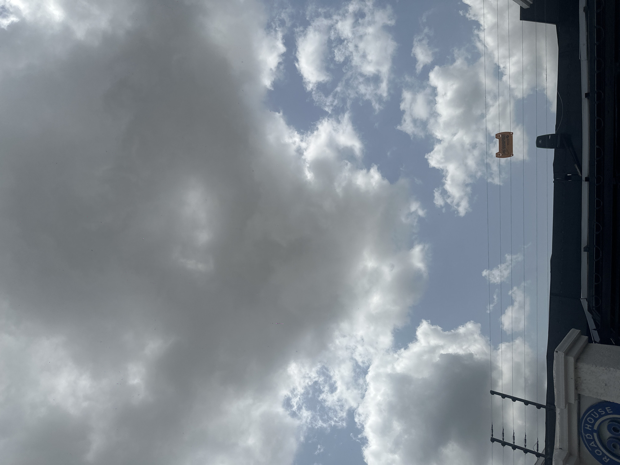 The image depicts a cloudy sky over a structure in Howayi, Nigeria. In the foreground, a white pillar with a blue circular emblem reading "ROAD HOUSE" is visible. Behind the pillar, a dark roofline extends across the image, with electric fence wires running horizontally above it. Suspended on these wires is a small orange object, possibly a marker or warning sign, with text that is difficult to read. The sky is a mix of dark, heavy clouds and patches of brighter blue sky with scattered white clouds.