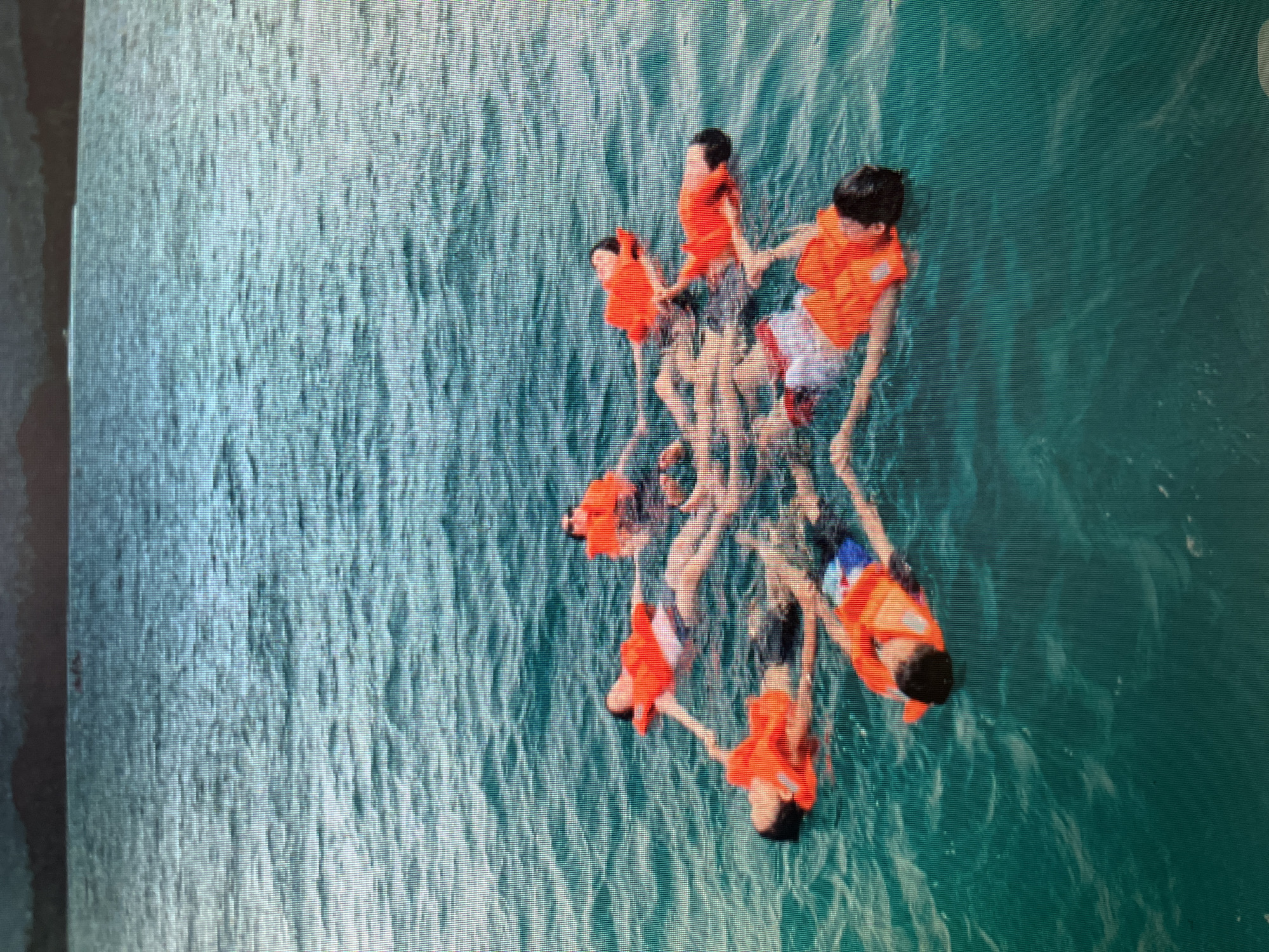 Eight individuals wearing bright orange life vests are floating on their backs in a serene turquoise body of water, forming a circular pattern. Their arms are extended outwards, with some individuals' hands touching or close to touching those next to them. The water is calm, with gentle ripples on the surface, and the background shows a dark, hazy shoreline with hills or mountains. The lighting suggests it is daytime, possibly late afternoon given the subdued colors of the distant land. The scene evokes a sense of relaxation and communal activity, likely a leisure pursuit in a coastal or lake area. The location is specified as Sejahtera, Indonesia, indicating a tropical or subtropical environment.