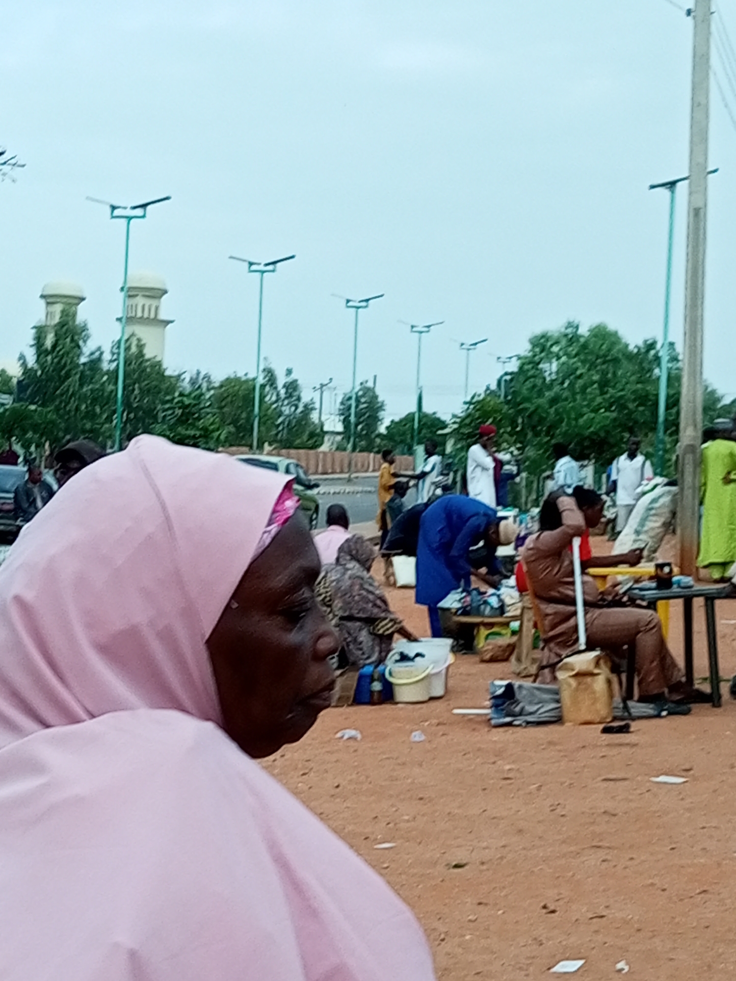 In Gombe, Nigeria, a crowded street scene unfolds under an overcast sky. In the foreground, a woman in a pink hijab is prominently featured in profile, her face partially obscured by the fabric and the angle of the shot. Behind her, other individuals are engaged in various activities. To the left, two figures, one in a dark jacket and another partially visible, stand near vehicles. In the middle ground, a gathering of people surrounds stalls laden with goods. One person in a blue robe bends over, appearing to tend to merchandise, while another in a brown outfit sits at a table, holding what seems to be a walking stick. A person in a bright green garment stands further back on the right. The environment is characterized by a dusty, reddish-brown ground littered with small pieces of paper. In the background, trees line a road, and prominent architectural features resembling minarets of a mosque are visible above the tree line, suggesting the proximity to a religious or significant public building. Several modern streetlights are also visible across the scene.