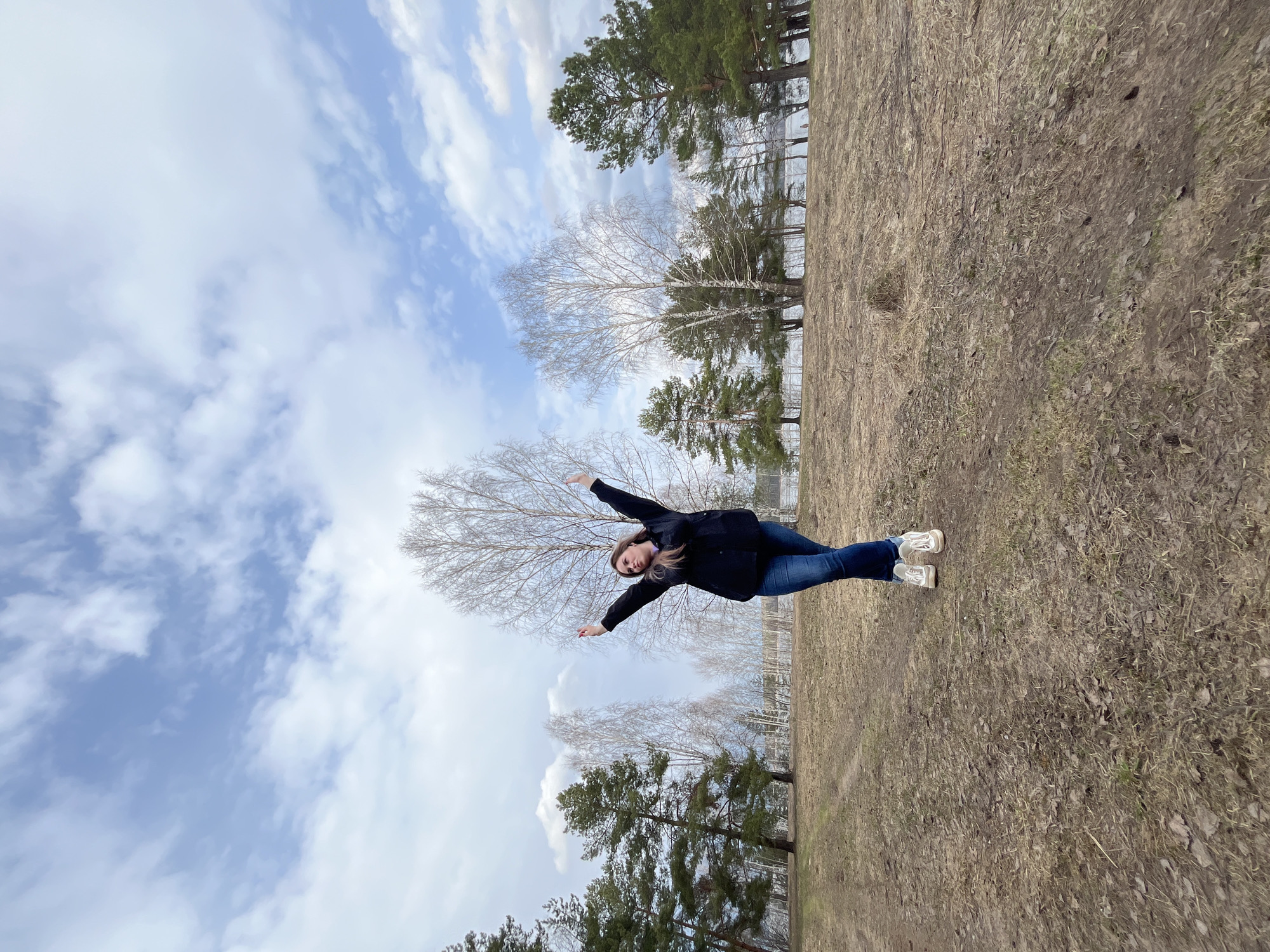 A woman is standing in a field with her arms outstretched, appearing to mimic an airplane. She is wearing a black jacket, blue jeans, and white sneakers. The background consists of a cloudy sky, trees, and a field of dry grass. The location is Chaikovsky, Russia.
