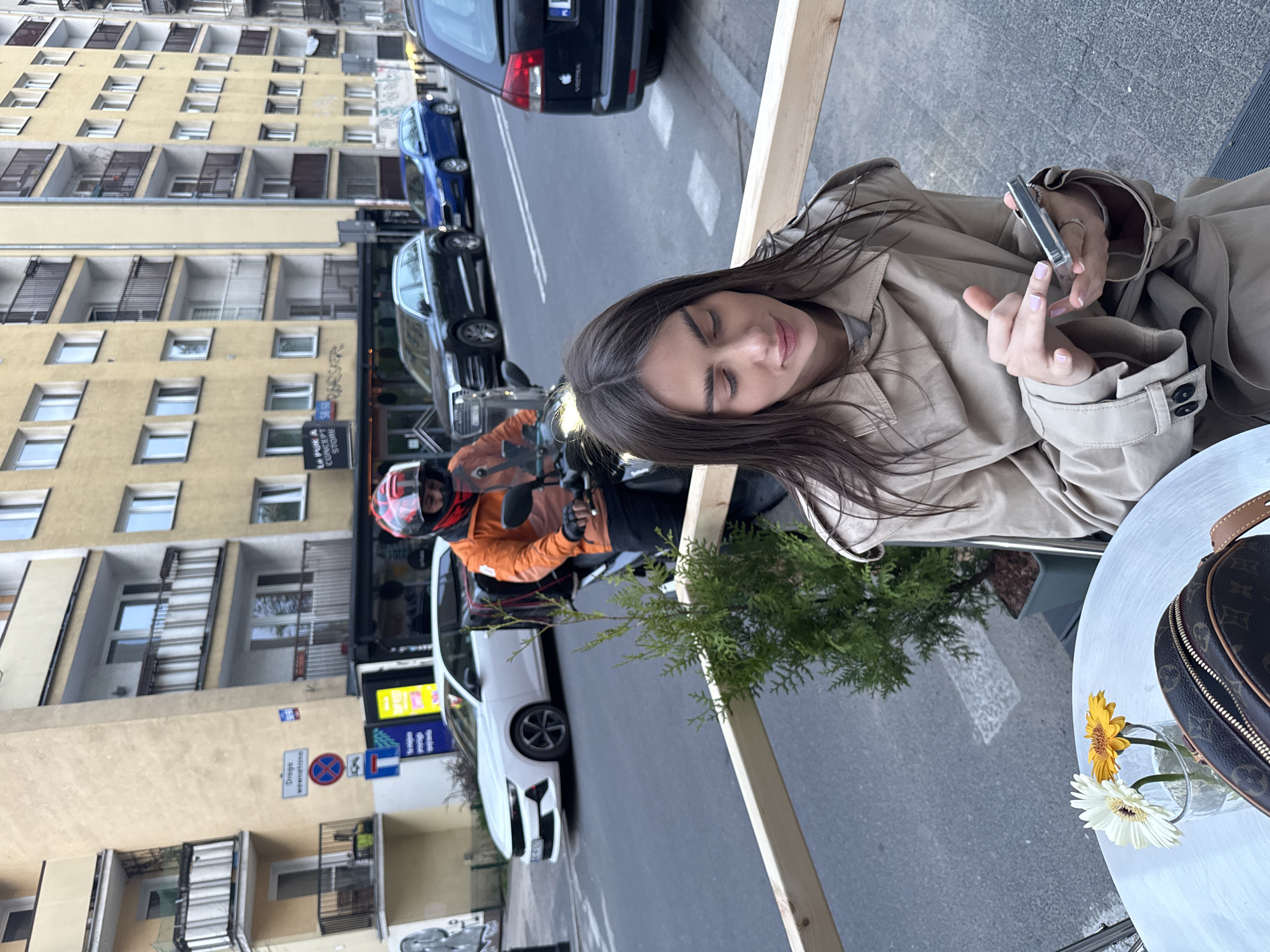 A woman with long brown hair, wearing a beige trench coat, is sitting at a white table on a city street. She is holding a silver smartphone. On the table, there is a small vase with two flowers, one yellow and one white, and a brown Louis Vuitton handbag. In the background, a man wearing a red helmet and orange jacket is on a motorcycle, next to a white car. The location appears to be in Warsaw, Poland, identifiable by the building architecture, parked cars, street signs reading "Droga Wewnętrzna" (Internal Road), and the "A PUKA CONCEPT STORE" sign visible on a store front. Other cars are parked along the street, and a tall yellow building with multiple windows is visible across the street.