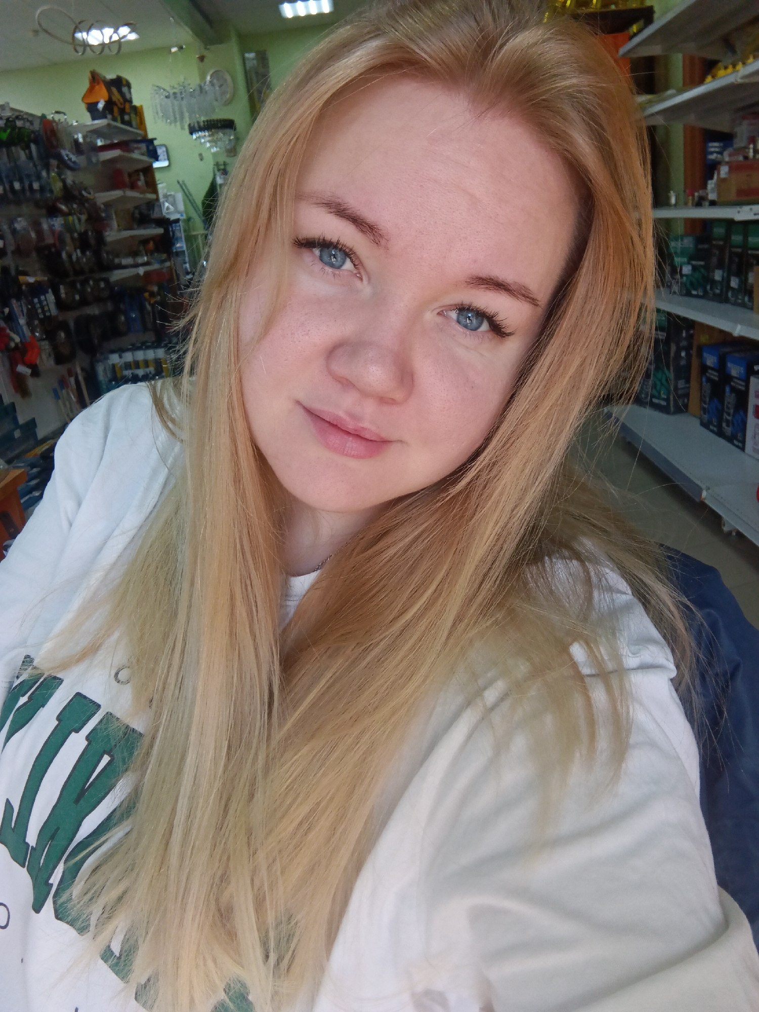 A young woman is featured in a close-up indoor selfie, likely taken in a retail or storage environment in Фоки, Russia. She has long, straight, light blonde hair that cascades around her shoulders and chest. Her blue eyes are directed towards the camera, and she has a subtle, pleasant smile. Her complexion is fair with visible freckles on her nose and cheeks. She is wearing a white long-sleeved top or t-shirt, which has a large green design or text, partially visible as block letters, on the left side of her chest.
The background indicates an indoor setting, with shelves filled with various products on both sides of the woman. On her left, light green walls are visible behind shelves stocked with what appear to be tools, hardware, or general merchandise. Decorative hanging items and a wall clock can also be seen. To her right, more shelves are lined with numerous items, possibly bottles or packaged goods. Overhead fluorescent lighting illuminates the scene. The overall impression is a casual, relaxed moment within a functional, commercial space.