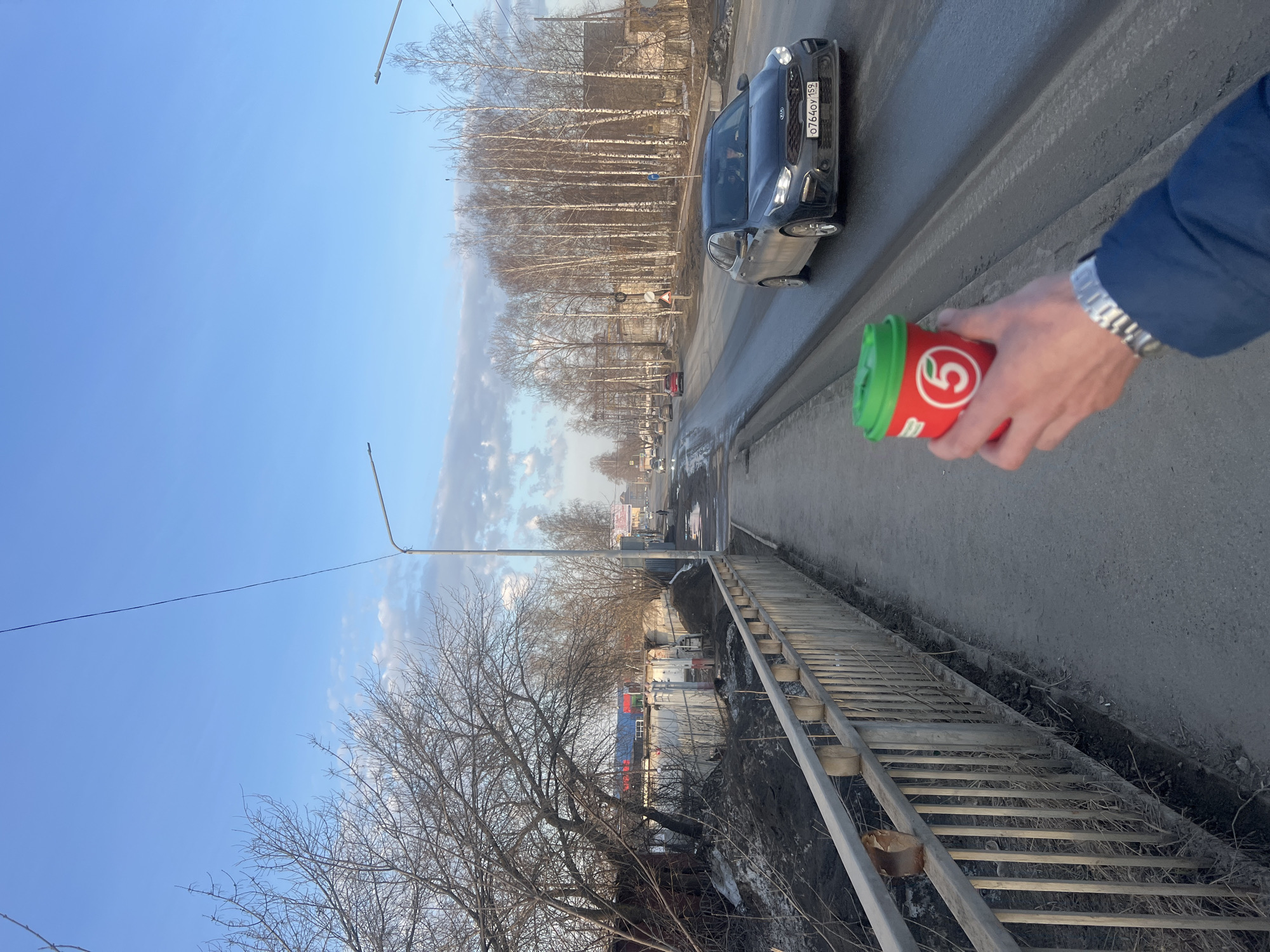 The image depicts an individual holding a coffee cup with the "5" logo of a Pyaterochka supermarket in Chaikovsky, Russia. The scene is set on a bridge with a metal railing overlooking a ditch with dry vegetation and residual snow. A road with vehicular traffic, including a dark-colored car with the license plate "O764OY159," runs adjacent to the bridge. Trees line the opposite side of the road, indicating an urban or suburban environment. The sky is clear and blue, suggesting daylight conditions. A street lamp is visible in the distance. The person holding the cup is wearing a blue jacket and a metal watch on their left wrist. The image appears to be taken from the perspective of someone walking along the bridge.