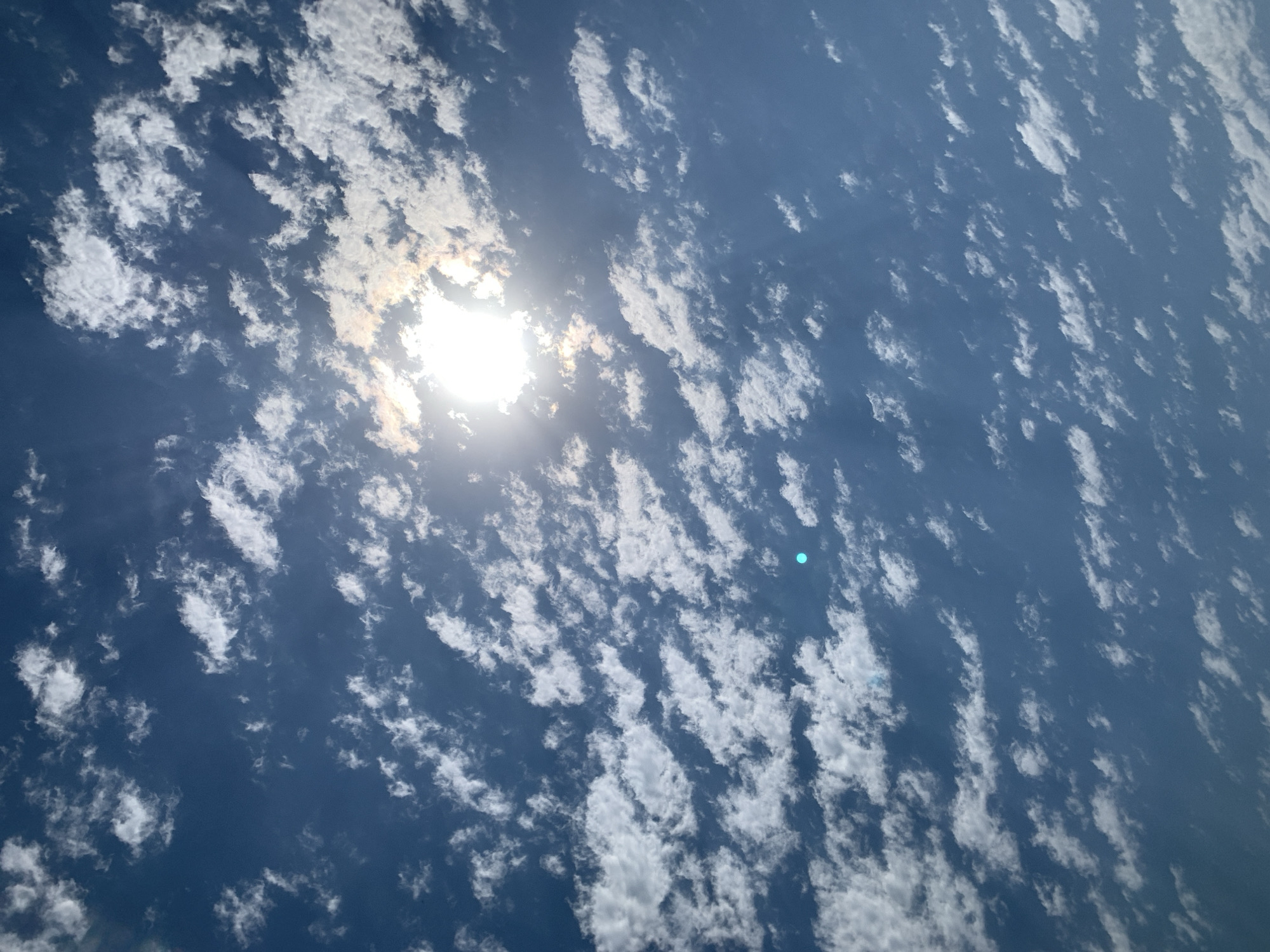 The image captures a bright, sunlit sky filled with scattered white clouds. The sun is prominently visible, partially obscured by clouds, casting rays of light. The sky is a deep blue. There are no people, structures, or discernible landmarks visible that would indicate a specific location in Khazar Raion, Azerbaijan. The scene is entirely focused on the atmospheric conditions.