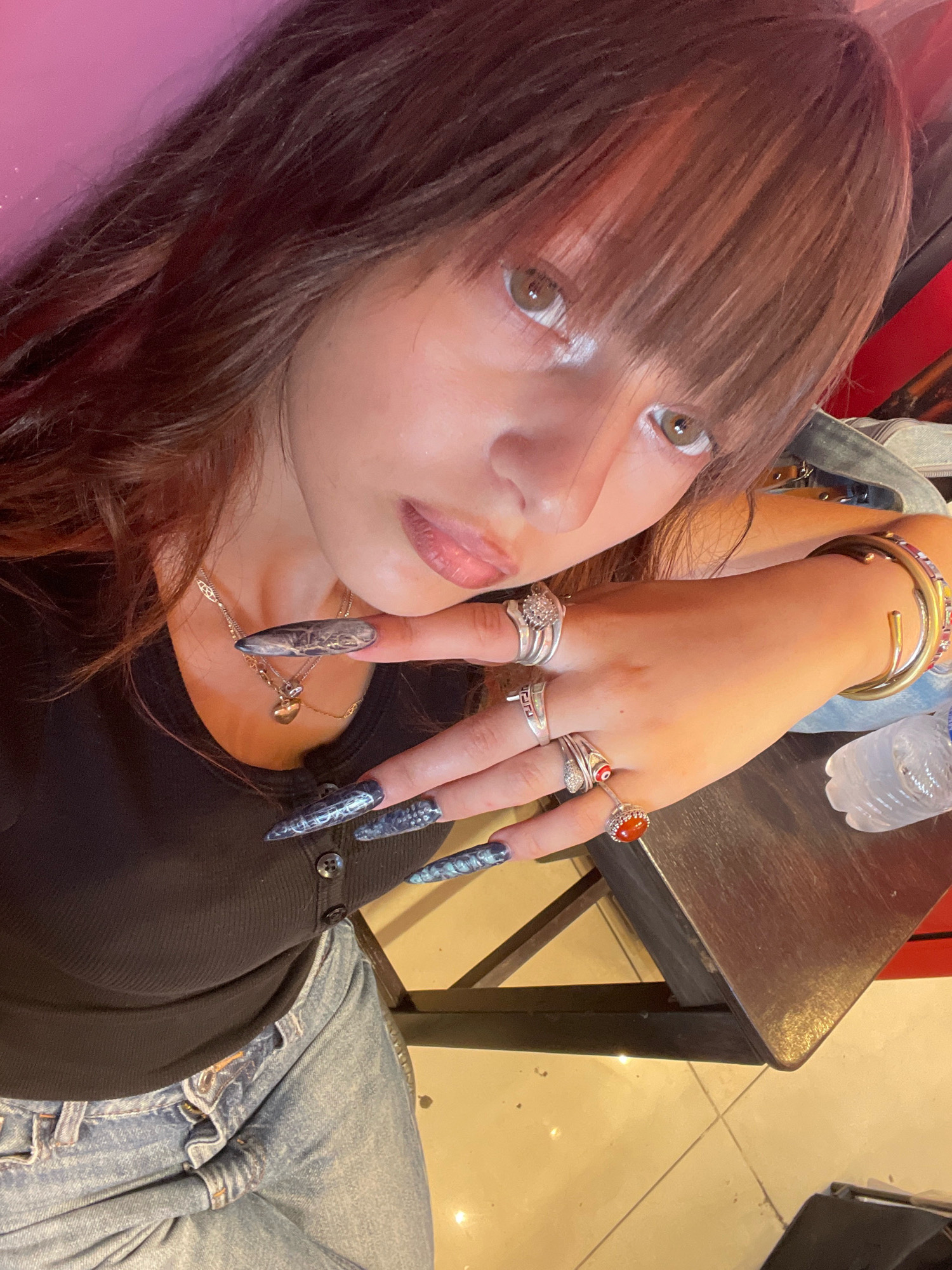 In this close-up, high-angle shot, a young person with long, dark brown hair and bangs is the central subject. They are wearing a black ribbed tank top with two visible buttons and light blue distressed jeans. The person has long, pointed nails adorned with a dark, patterned design, possibly resembling snakeskin. They are wearing several rings on their fingers, including one with a red stone and another with a cluster of small stones. A gold necklace with a heart-shaped pendant is visible around their neck. They are also wearing a thick gold bangle on their wrist.
The person is leaning on a dark wooden surface, possibly a table. A partially visible plastic water bottle with a white label is also on the surface. The background is a mix of pink and red tones, with some blurred elements that suggest an indoor setting. The lighting appears to be warm and artificial, casting shadows on the table and floor. The person's gaze is directed towards the camera, with a neutral expression. The overall impression is a selfie taken in a casual, possibly indoor environment. There is no visible text in the image. The location context provided is Raanana, Israel.