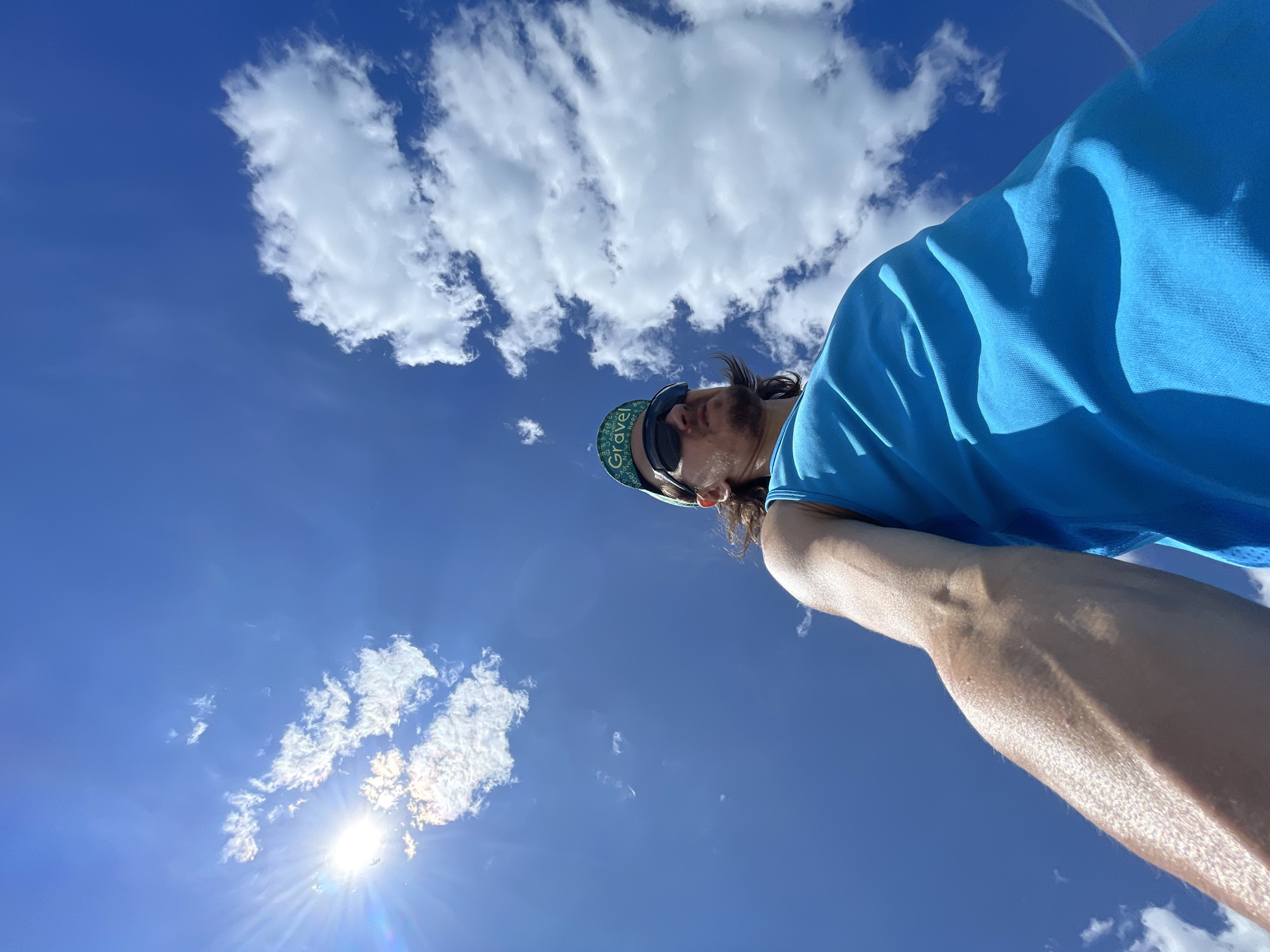 A low-angle, upward-facing self-portrait features a male individual against a clear, vibrant blue sky dotted with white cumulus clouds. The subject, positioned in the lower portion of the frame, wears a bright blue sleeveless top, dark wrap-around athletic-style sunglasses, and a green baseball cap with the word "Gravel" prominently displayed in white text on the front. His face is turned slightly to the viewer's right, with a chin-length beard visible, and longer hair flowing from beneath the cap. His right arm (viewer's left) extends towards the camera in the bottom-left corner, showcasing a muscular forearm with visible veins. The sun, emitting a strong lens flare and starburst effect, is visible in the upper-left quadrant of the sky, casting bright light on the scene. The scattered white clouds range from small wisps on the left to larger, puffier formations on the right. The overall impression is a bright, sunny day outdoors.