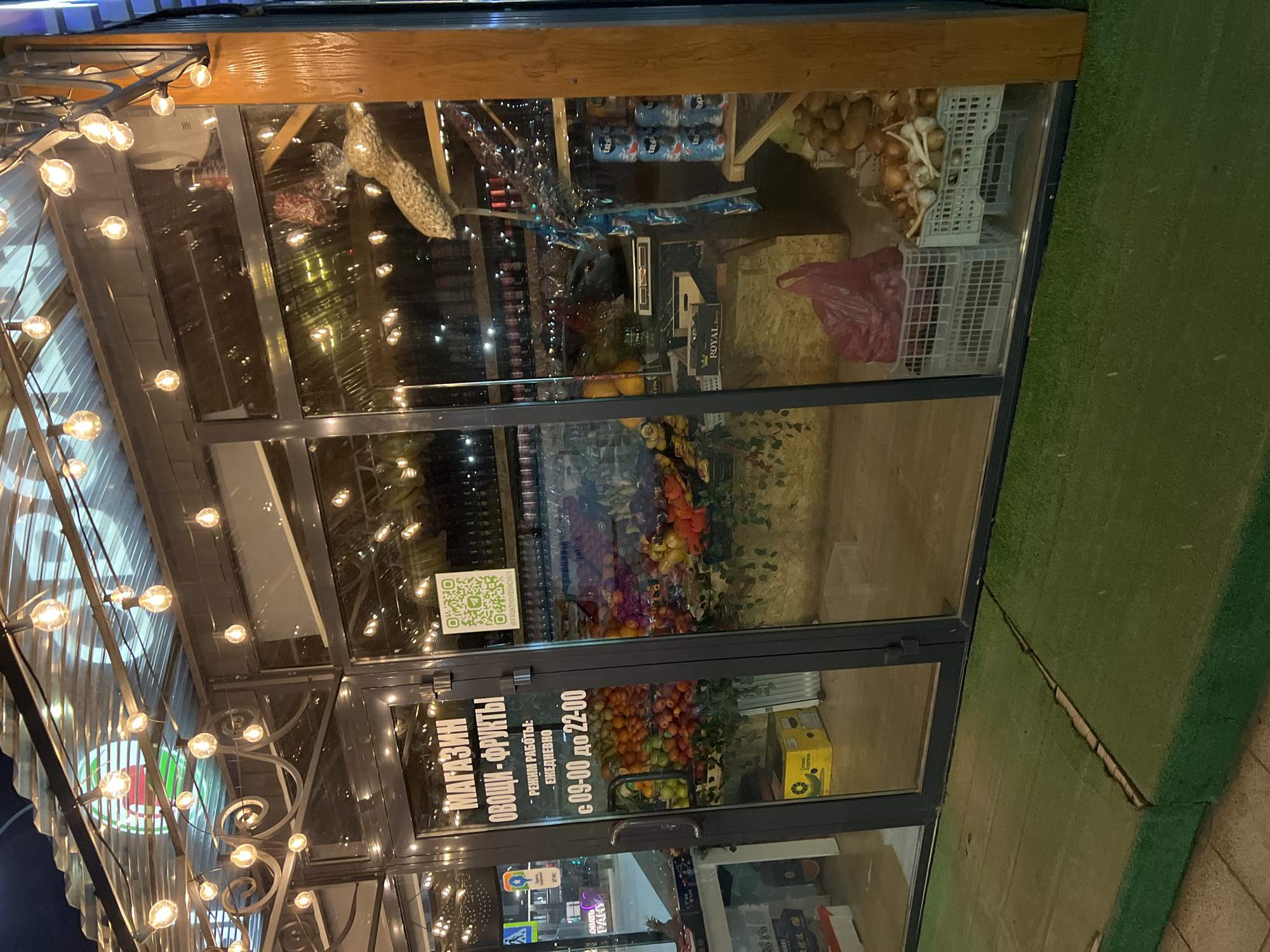This image shows the exterior of a fruit and vegetable shop in Tyumen, Russia. The storefront is glass, and the sign on the door reads "МАГАЗИН ОВОЩИ - ФРУКТЫ", which translates to "Shop Vegetables - Fruits". The shop's hours are listed as daily from 09:00 to 22:00. Inside, shelves are stocked with various fruits, vegetables, and canned goods. Outside, decorative string lights illuminate the entrance. The area adjacent to the shop is covered in artificial turf. A QR code sticker is visible on the door. Reflections in the glass show the surrounding urban environment.