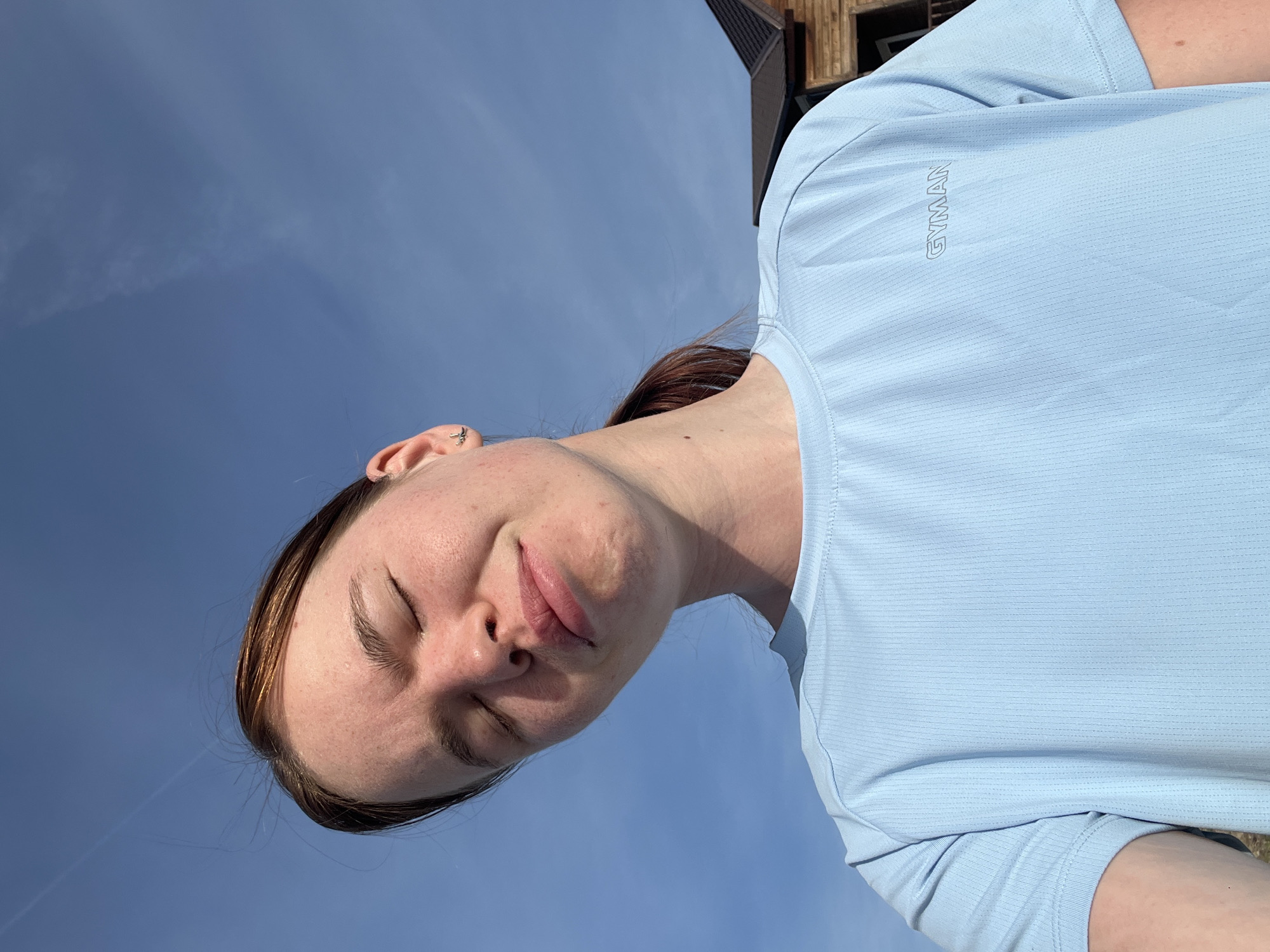 A young fair-skinned woman is angled upwards towards a blue sky. The woman is wearing a light blue t-shirt with the word "GYMAN" printed on the right side of the shirt. The building behind the woman suggests she may be in Khvalynsk, Russia. Her eyes are closed and she has brown hair tied back.