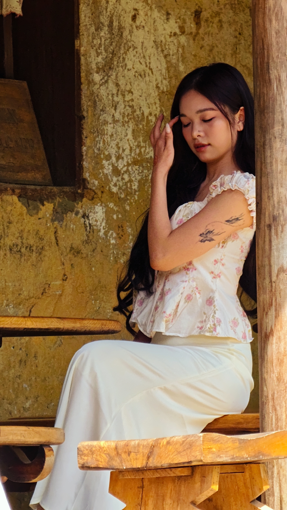 A young Asian woman with long dark hair is sitting on a wooden bench in front of a textured, weathered yellow wall. She wears a white floral top and white skirt. Her right hand is raised near her forehead. A tattoo of three birds in flight is visible on her upper left arm. The background suggests a rustic setting, possibly in Lũng Cú Commune, Vietnam, given the architectural style and environment. The lighting indicates an outdoor daytime scene. The woman is situated on a wooden structure that appears to be part of a building or sheltered area.
