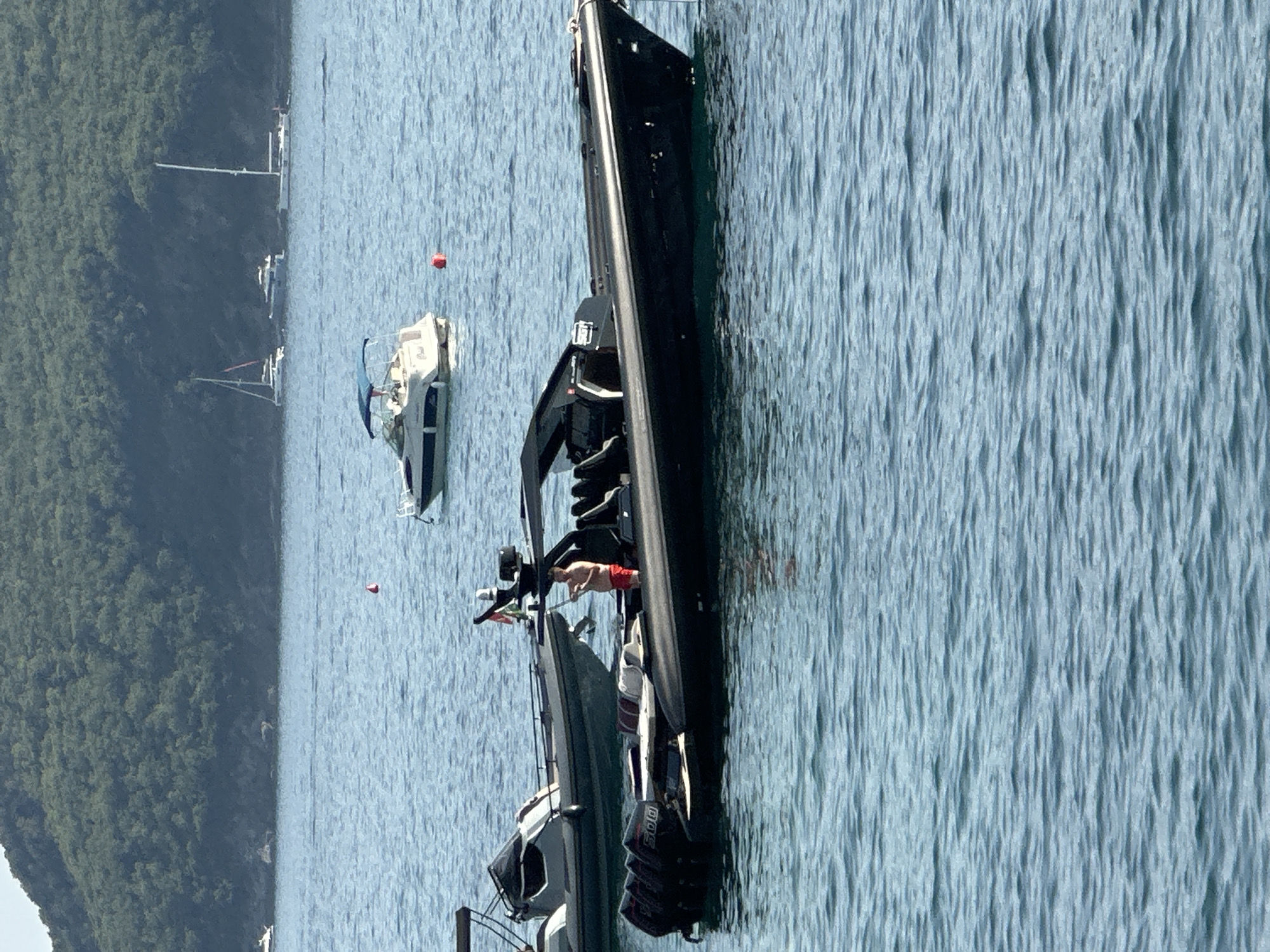 The image depicts a sunny day at sea, with several boats anchored near a lush, green hillside. In the foreground, a sleek, black speedboat is prominently featured, with a man in red swim trunks standing on it, holding a pole. Behind this boat, another gray speedboat is visible. In the middle ground, a white motorboat with a blue canopy is anchored, and further in the distance, several sailboats and other vessels dot the water. The background shows a densely forested hillside that slopes down to the water, characteristic of the Budva Riviera in Montenegro. The water is a calm, blue expanse, with gentle ripples on the surface.
