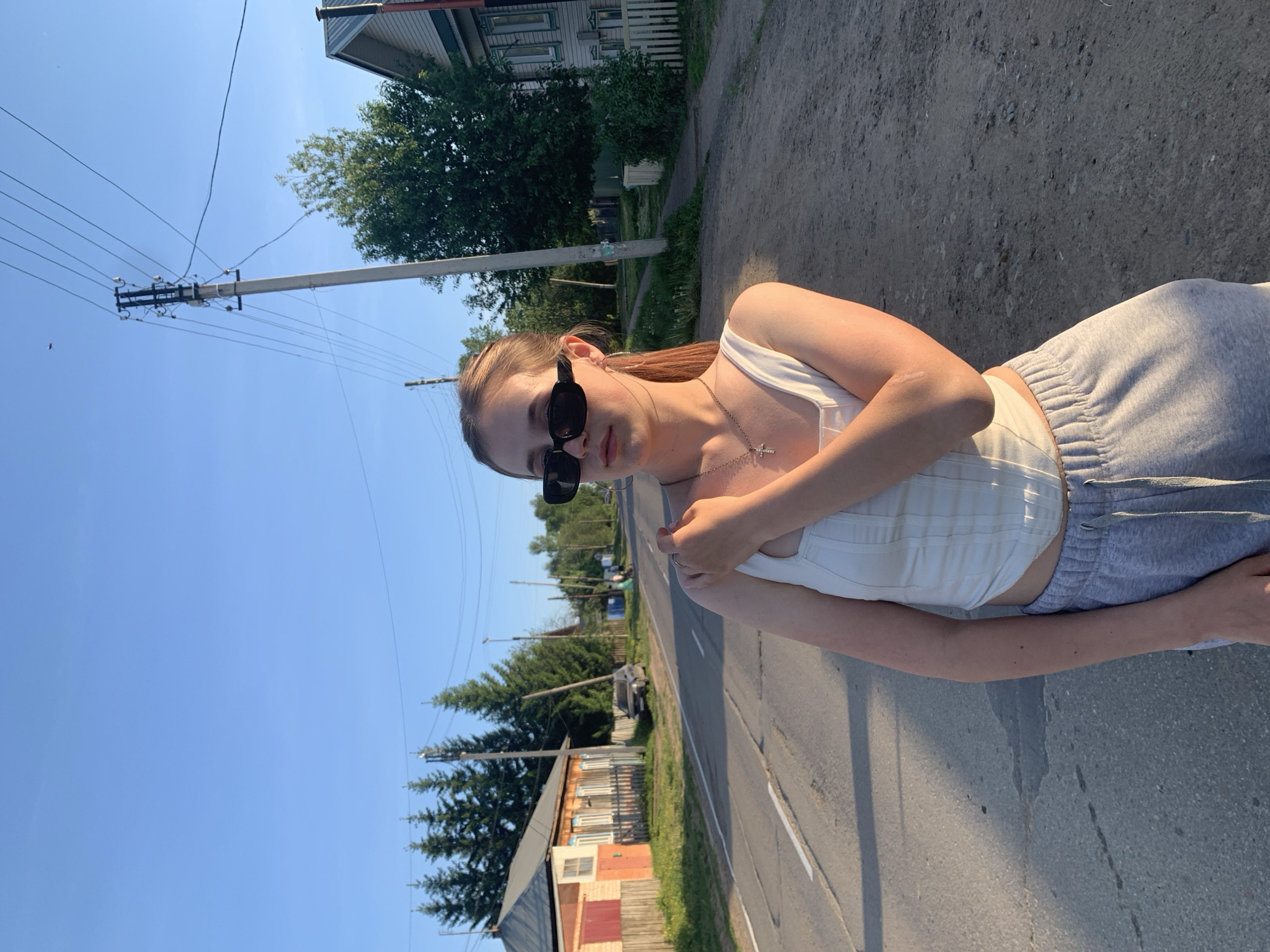 A young woman stands on a gray asphalt road, in what appears to be the city of Фоки, Russia, indicated by the presence of typical residential buildings and power lines. She wears dark sunglasses, a white sleeveless top, light gray sweatpants, and a silver necklace with a cross pendant. The area is bathed in sunlight under a clear blue sky. Behind her are houses, trees, and utility poles, giving context to a small-town street setting.