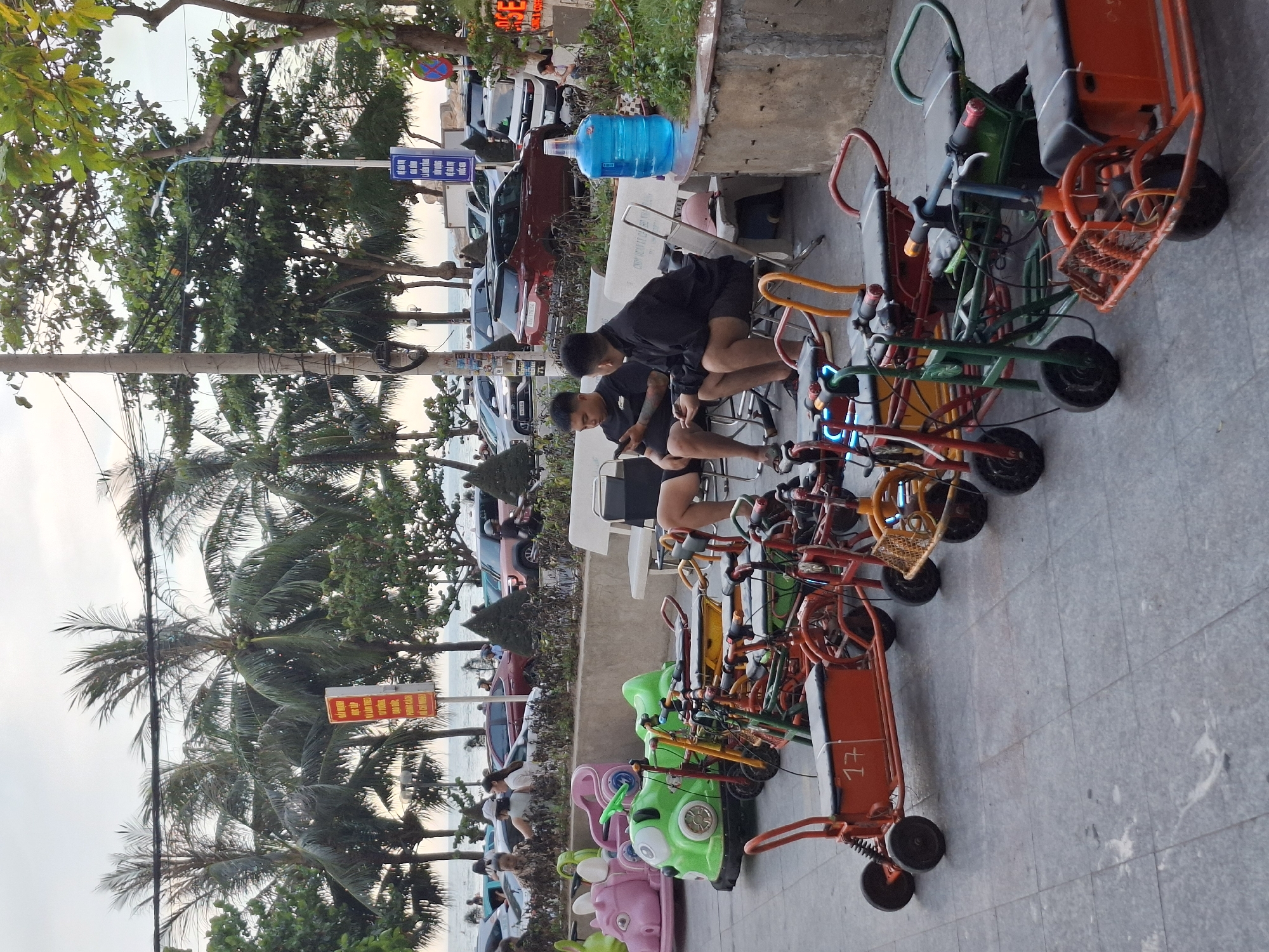 The image depicts a street scene in Bac Nha Trang Ward, Vietnam. The primary subjects are several people and a collection of small, colorful vehicles, likely for rent. Two young men in black shirts are seated on chairs, one looking at his phone. In the background, a row of parked cars suggests a public area.
The scene is filled with an assortment of small, child-sized vehicles. These include bright green and pink "cartoon" themed rides with playful designs, and more traditional red and orange pedal carts with baskets and seats. They are lined up on a paved sidewalk, suggesting a rental or amusement area.
Behind the vehicles, palm trees and other lush greenery are visible, with the ocean hinted at in the distance. The sky appears overcast, suggesting a possibly humid or cloudy day, typical of coastal tropical climates.
Visible text includes a blue sign with white lettering on a pole, and a red and yellow sign on a pole near the palm trees with Vietnamese text. One of the red carts also has the number "17" clearly marked on it.
The overall impression is a relaxed, casual atmosphere, possibly a tourist area where families might rent these small vehicles for entertainment. The presence of parked cars and the general street setting place the scene in an urban or semi-urban environment, likely near a beach or promenade.