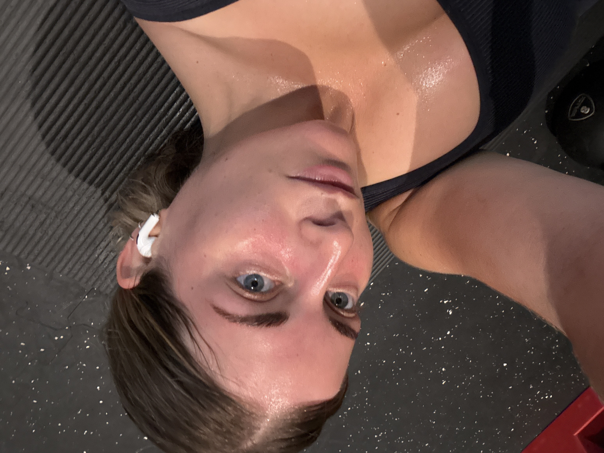 A close-up, upside-down view captures a woman lying on a textured, possibly rubberized, surface. Her face, flushed and glistening with sweat, suggests recent exertion. She has fair skin, light blue eyes, and dark eyebrows. Her lips are slightly parted. A white wireless earbud is visible in her left ear. She is wearing a dark-colored, possibly ribbed, sports bra. The background includes glimpses of the textured surface she is lying on and part of a black object. The lighting appears to be artificial and somewhat harsh, highlighting the sheen of perspiration. No indication of location in Paris.