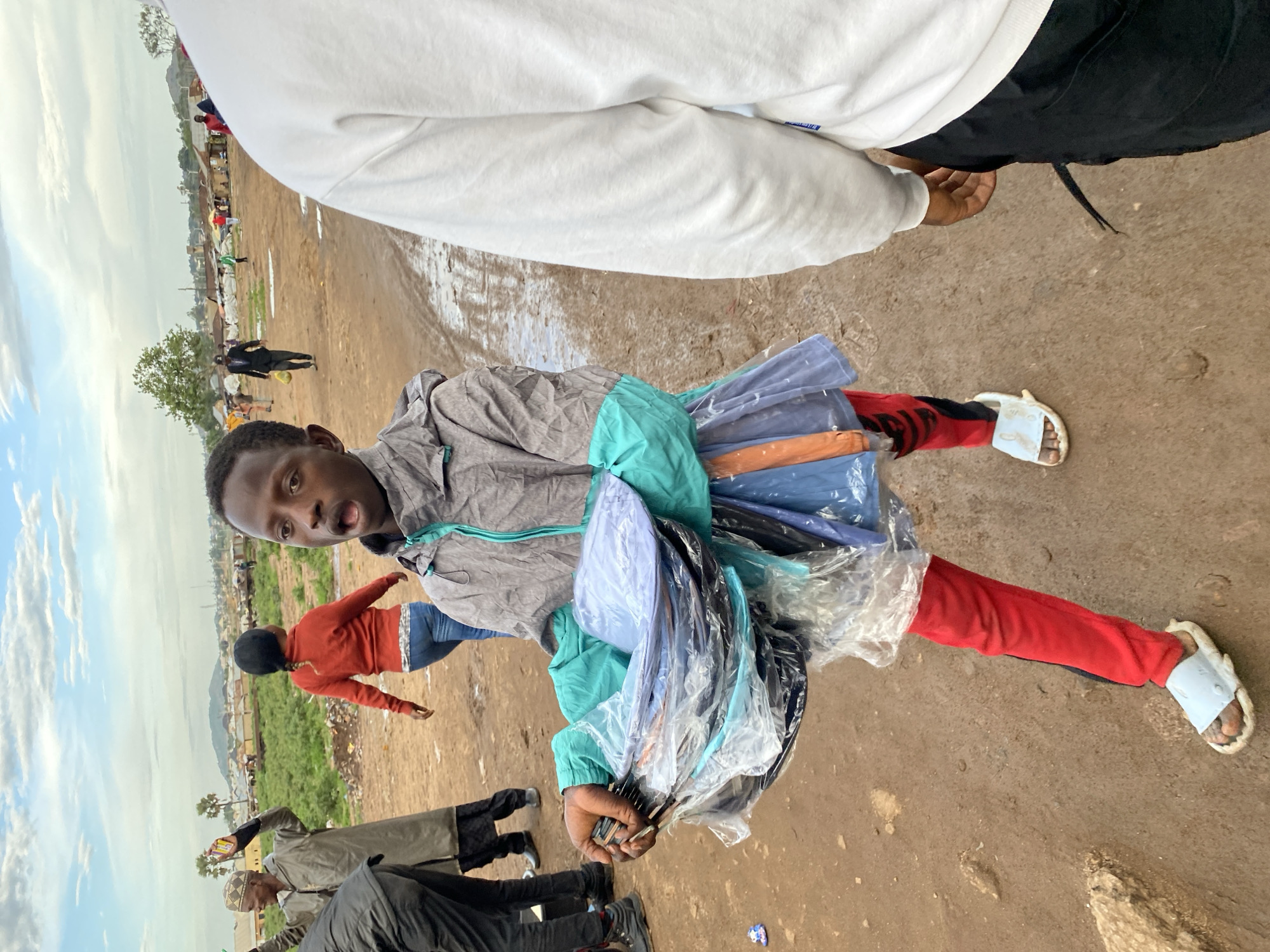 The image depicts an outdoor market or street scene in Bukuru, Nigeria, under an overcast sky.
In the immediate foreground, a young male, appearing to be a hawker, stands center-left, facing slightly right of the viewer but looking directly at the camera. He wears a grey and teal two-tone hooded jacket, red pants with black vertical accents on the outer seams, and light blue slide sandals over bare, dirt-covered feet. His mouth is open, and his eyes are wide. He carries a substantial bundle of neatly folded clothing items, individually wrapped in clear plastic bags, in his arms at waist level. The clothing items are visible in various solid colors, including blue, black, light blue, and orange.
To the right of the hawker, the partial back of an individual wearing a white long-sleeved sweatshirt and black pants is visible, extending from the right edge of the frame to the mid-right.
In the left midground, an older male in a light-colored traditional robe (agbada) and a patterned cap stands with both arms raised, holding small yellow objects in his right hand. He faces away from the camera, looking towards the left. Behind him, another individual, possibly a woman, in a red long-sleeved top and blue jeans, with dark hair or head covering, walks away from the viewer towards the left.
Further in the midground, towards the center-left, a man in a dark suit jacket and pants, possibly a dark hat, walks towards the left, carrying a yellow bag in his left hand. Additional indistinct figures are scattered throughout the background.
The ground is predominantly reddish-brown dirt, uneven, with small visible rocks and patches of mud, particularly a muddy track extending from the midground towards the background on the right. Sparse green vegetation and a few trees, including two palm trees on the left and a larger deciduous tree in the center, are present in the midground and background. Distant, low-rise buildings with light-colored roofs and some makeshift structures are visible on the horizon, indicating a populated area. The sky is largely cloudy with patches of blue.