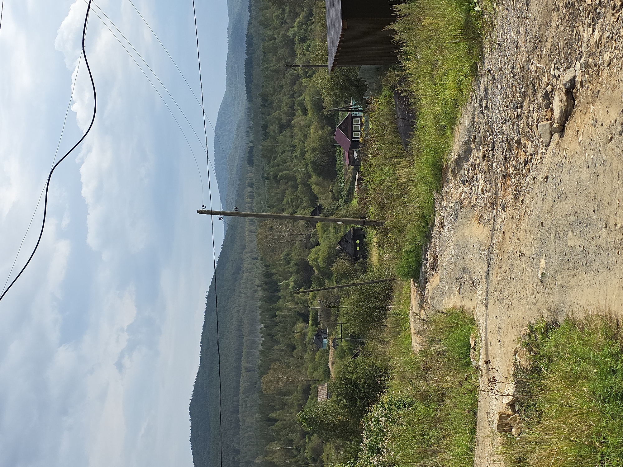 The image depicts a rural scene with a dirt road leading through a landscape of rolling hills covered in dense green forest. The sky is overcast with scattered clouds. In the mid-ground, several small, rustic houses with wooden structures and pitched roofs are visible amidst trees and vegetation. A wooden utility pole stands along the road, and power lines stretch across the upper portion of the frame. The foreground shows the unpaved road with loose gravel and some rocks, flanked by tall grasses and wild vegetation. The overall impression is one of a remote, undeveloped settlement within a natural, forested environment. The location is identified as Magnitka, Russia.